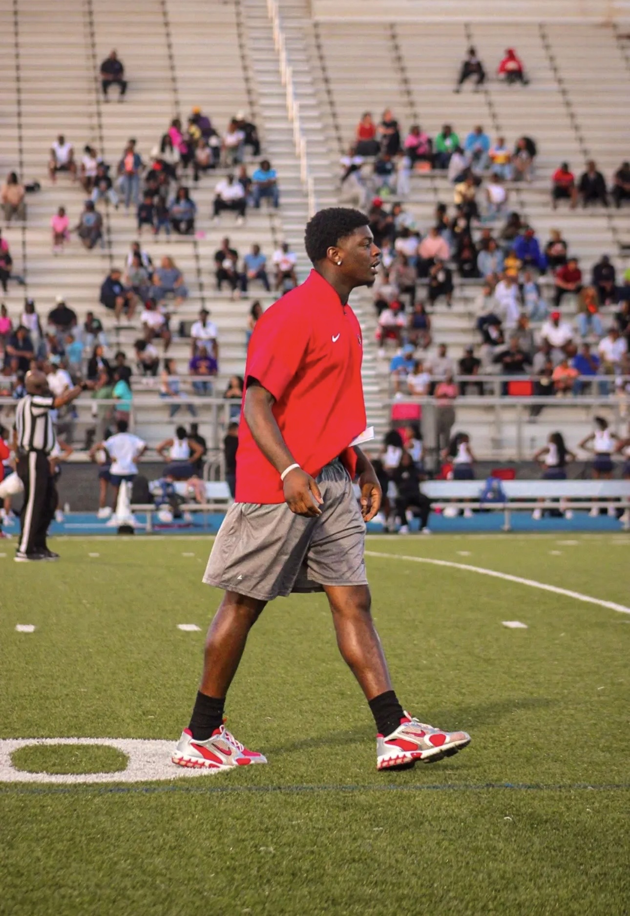 Young man walking on a sports field with spectators in the background, wearing a red shirt and gray shorts.