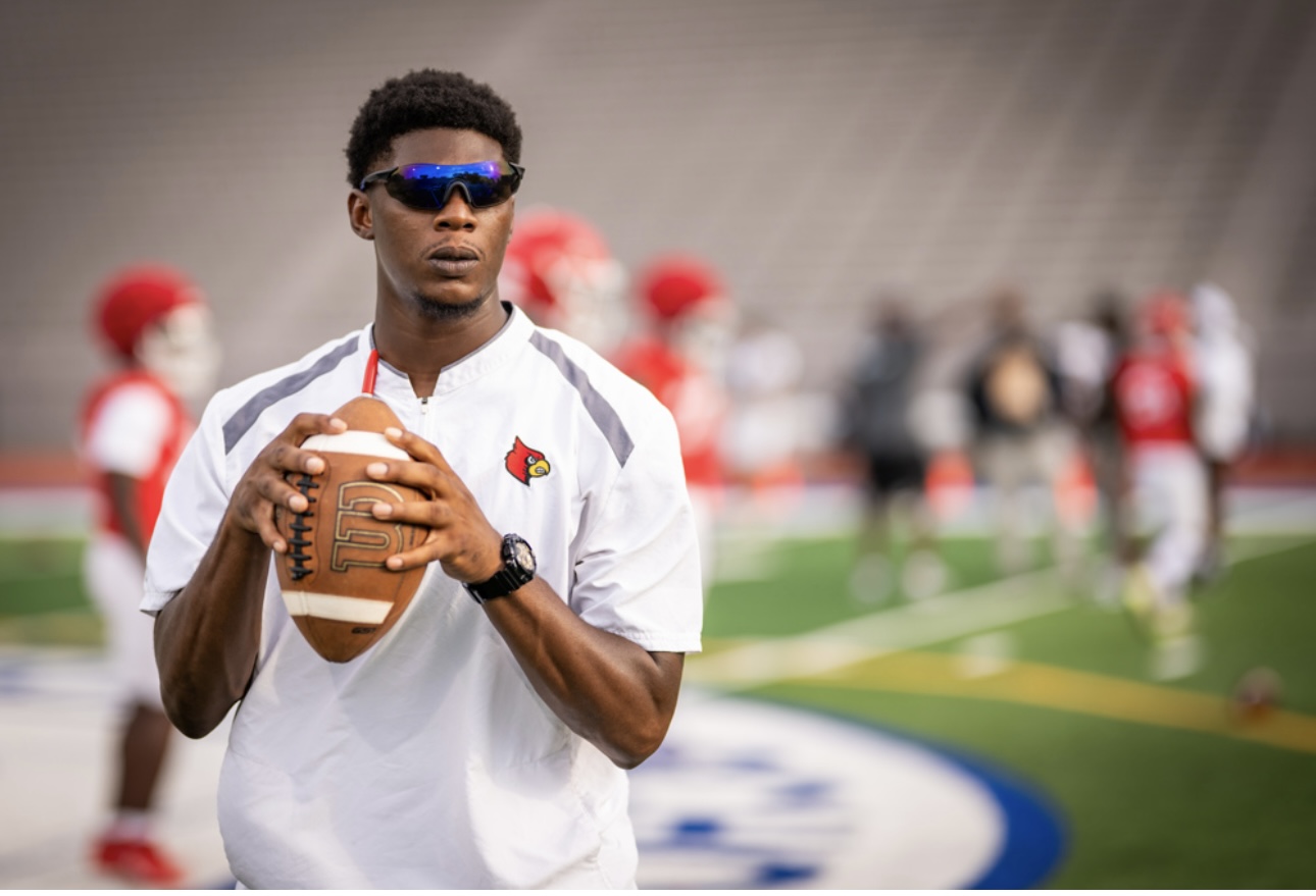 Young man in sunglasses holding a football on a sports field with players and spectators in the background.