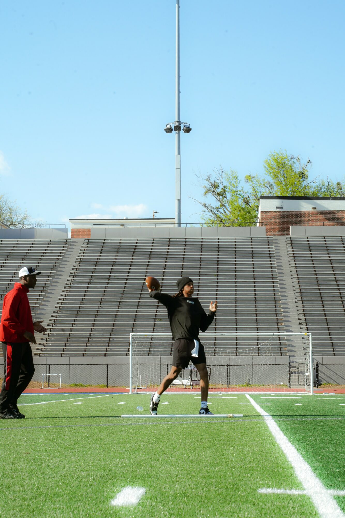 Two people on a sports field, one holding a ball, with a stadium and trees in the background.