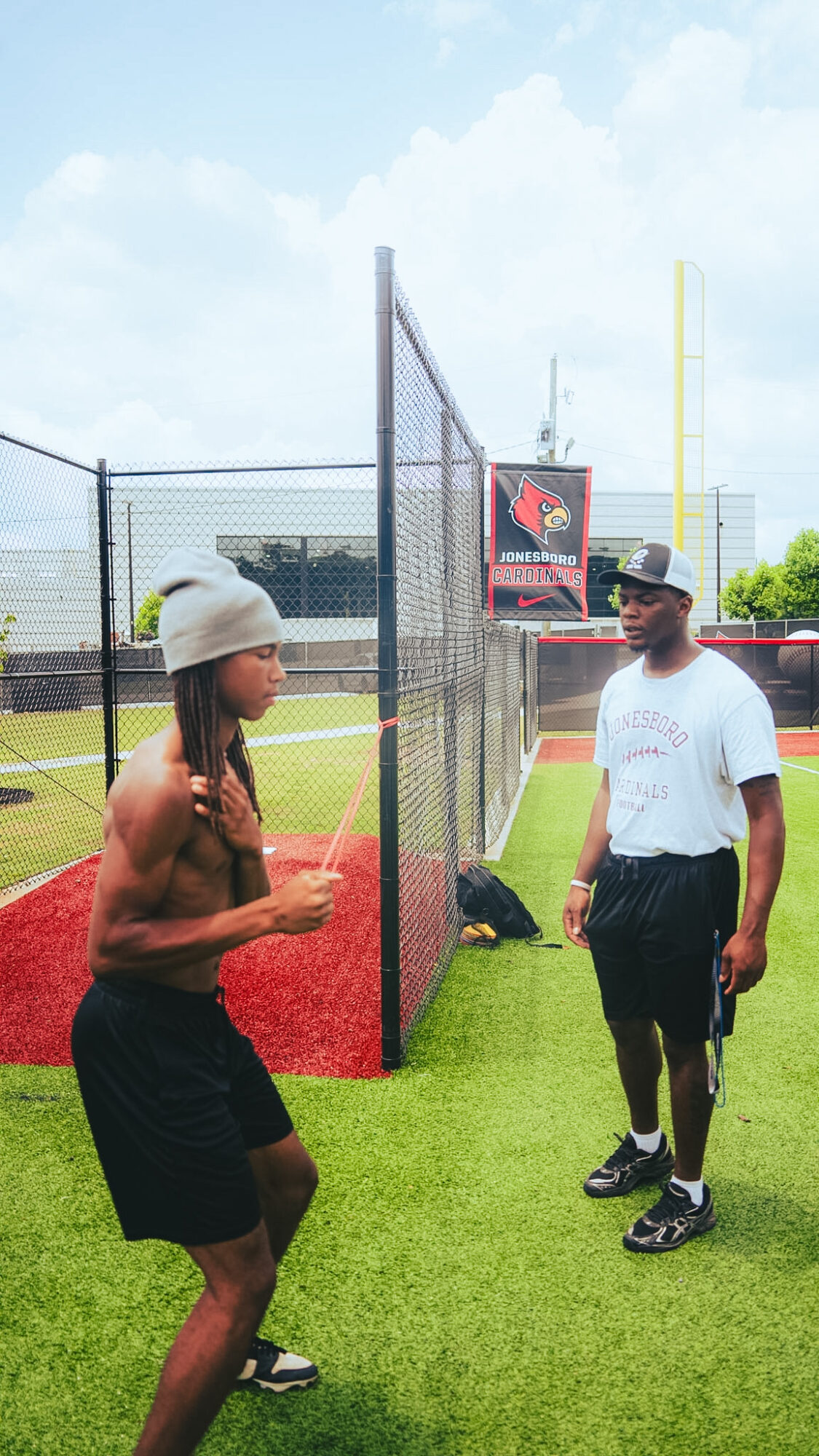Two men talking on a sports field with a fence and scoreboard in the background.