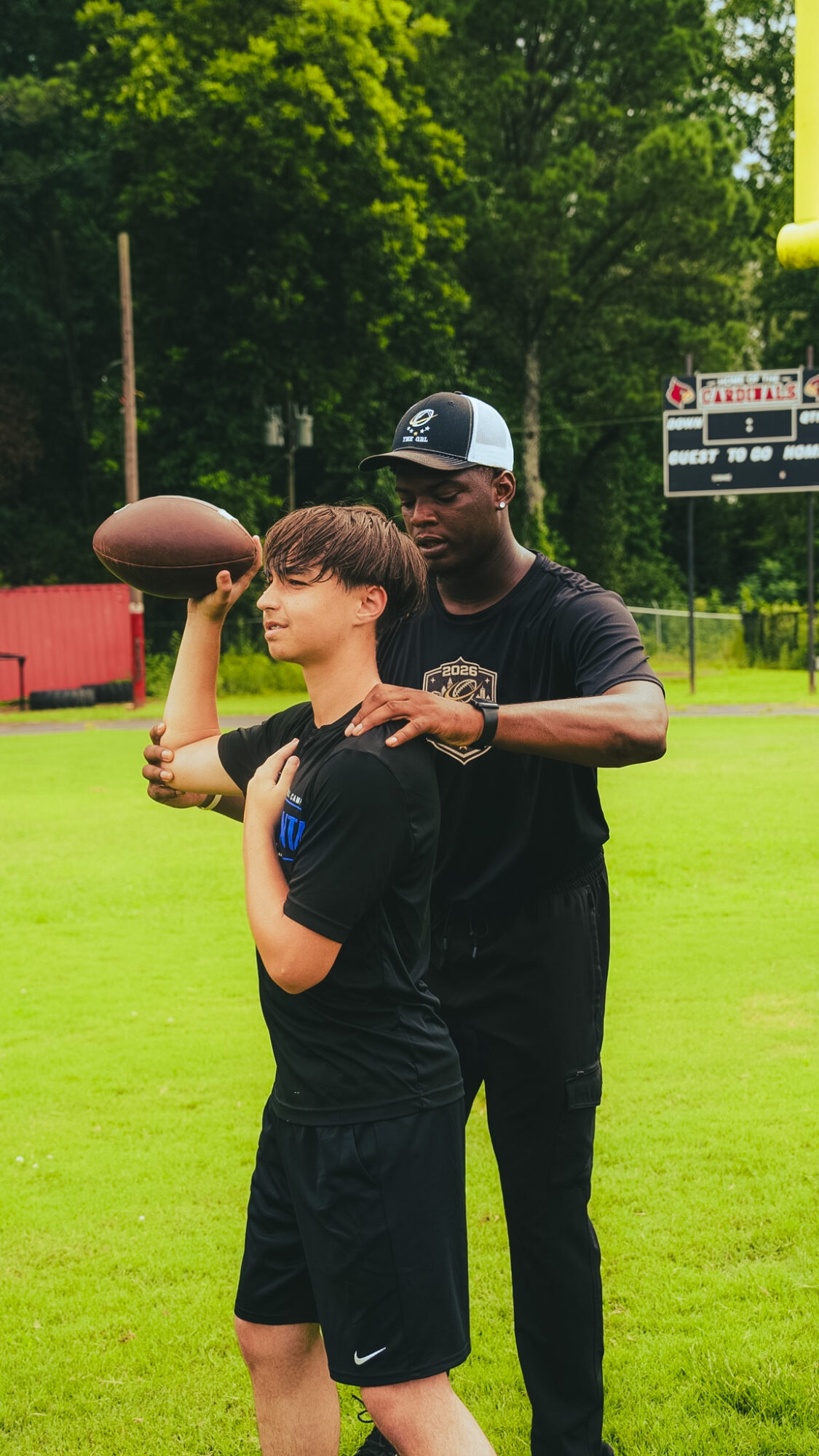 Two people on a grassy field, one holding a football, the other guiding or coaching, with trees and a scoreboard in background.