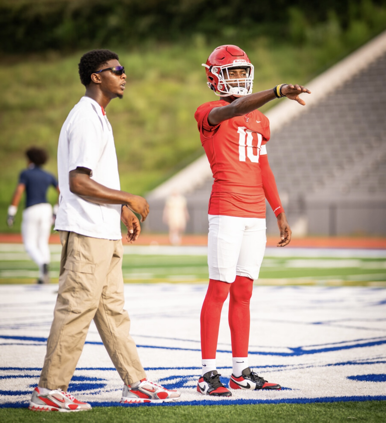 Football player in red uniform points on the field, beside a person in casual clothing, on a stadium field.