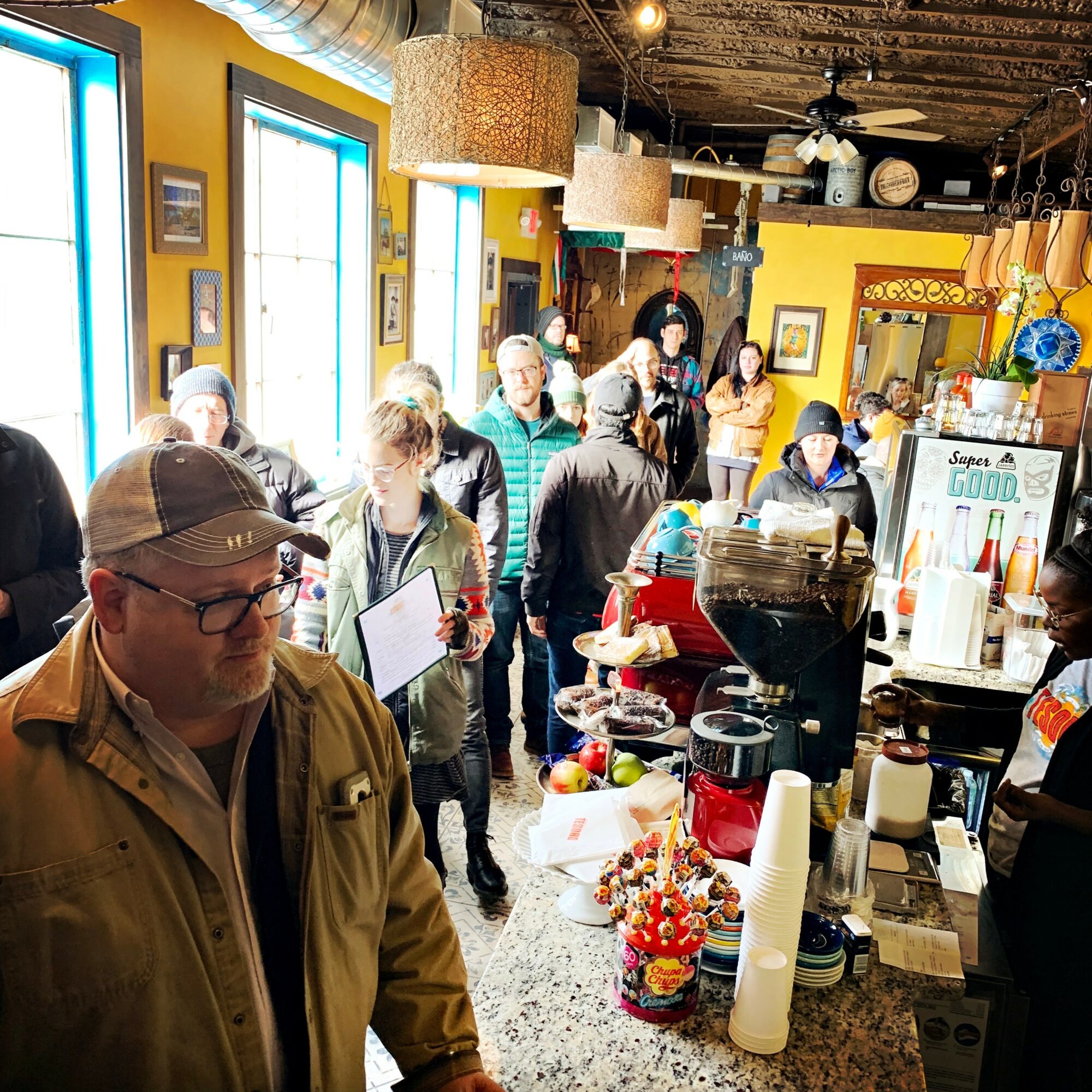 People gather inside a brightly lit cafe with yellow walls, large windows, and various decorations, some wearing masks.