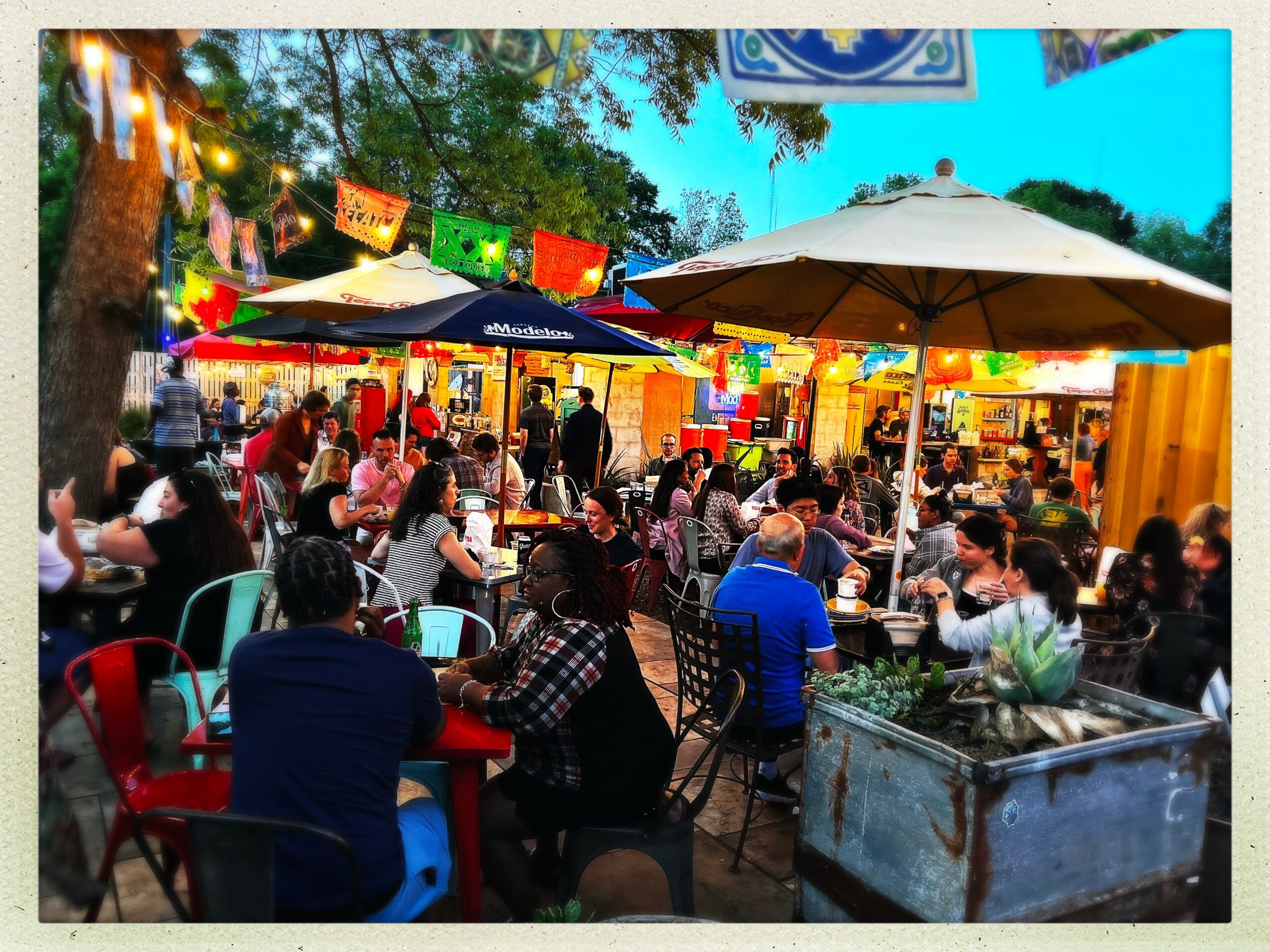 Outdoor market with people sitting at tables, colorful banners, umbrellas, and food stalls, surrounded by trees and string lights.