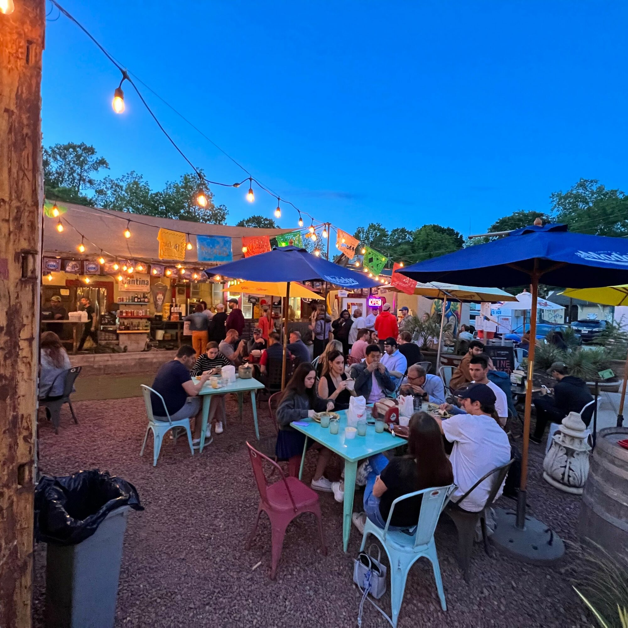 People dining outdoors at tables with umbrellas under string lights during evening, lively atmosphere, food stalls in background.