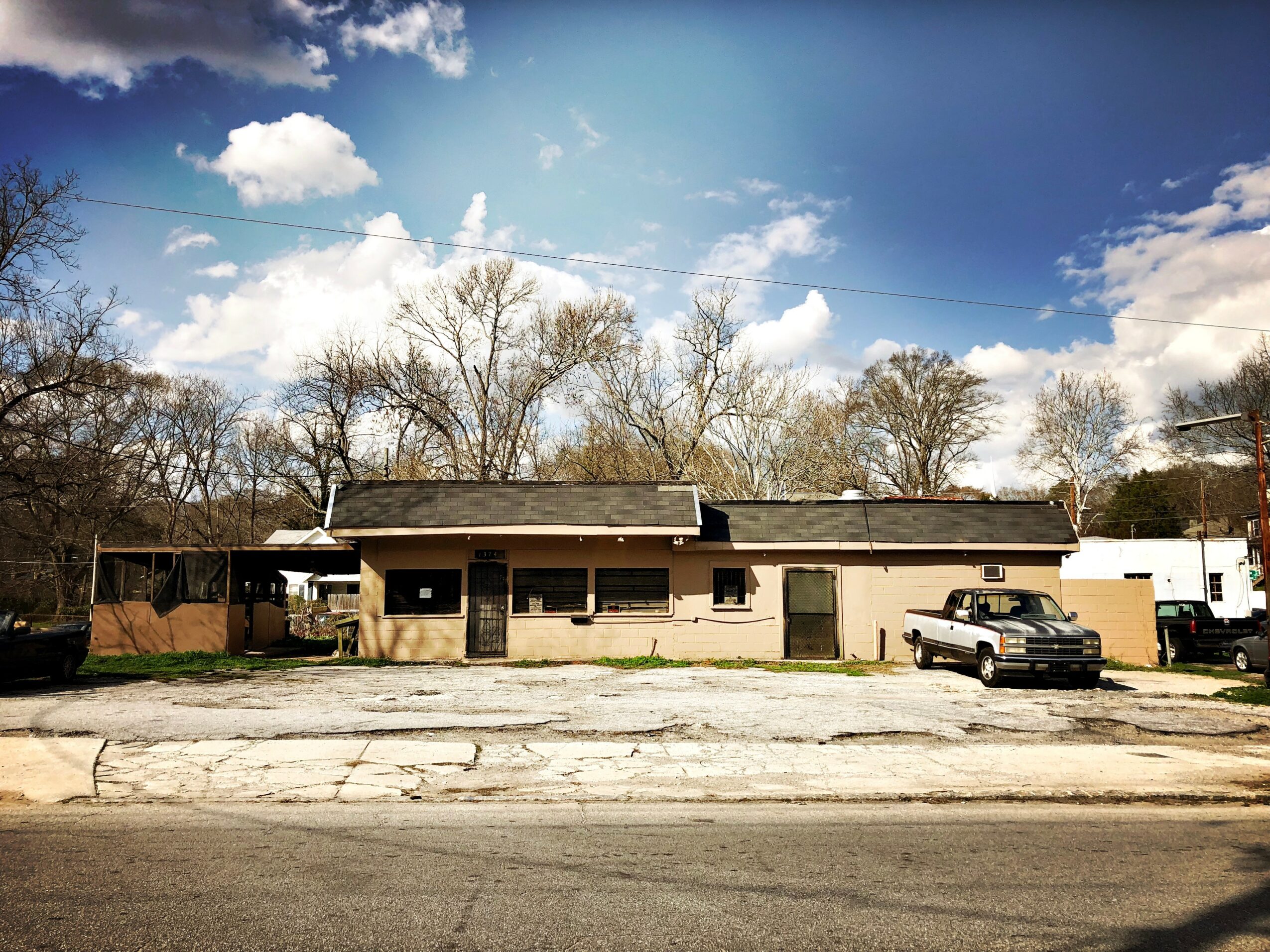 Single-story building with trees and a partly cloudy sky in the background, parked car in front, and a cracked pavement in the foreground.