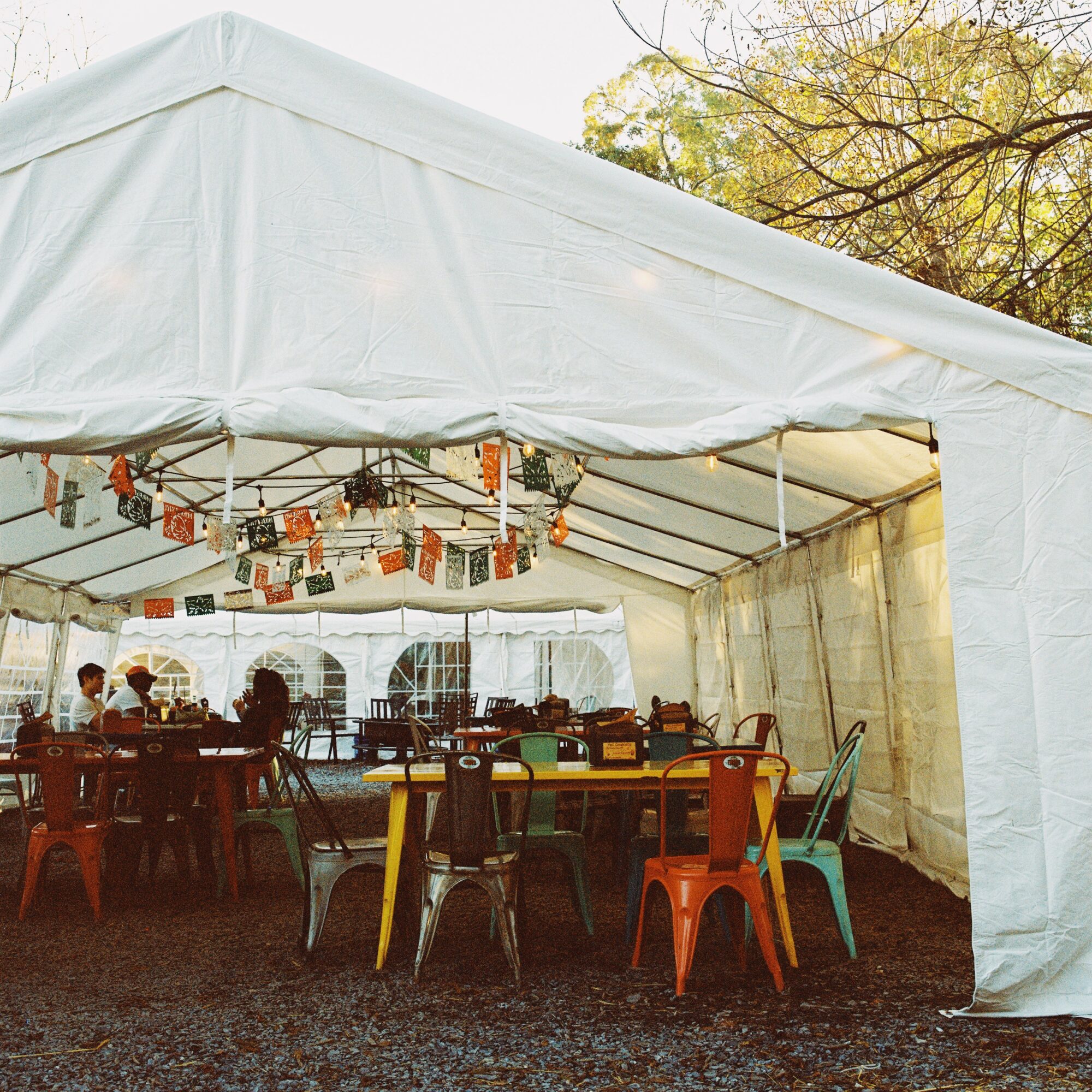 Inside a large white tent with colorful chairs and tables, some people sitting, hanging decorations, and trees outside.