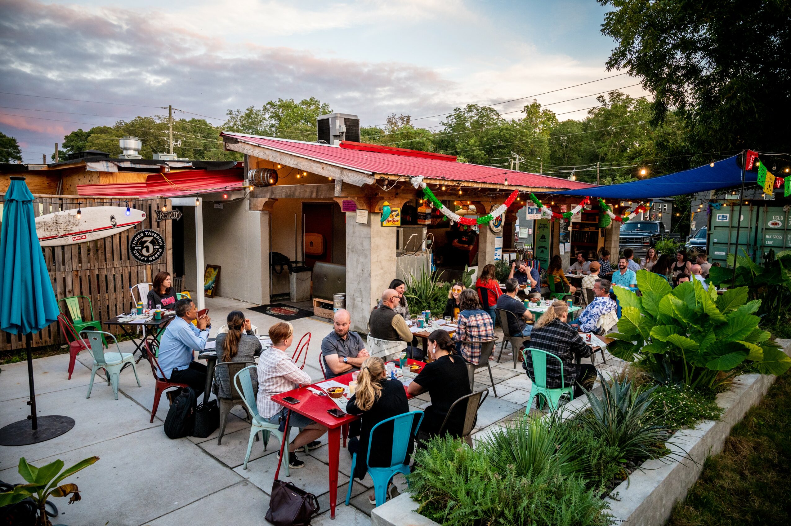 People dining outdoors at tables with colorful umbrellas and decorations, in front of a building with a red awning, trees, and a cloudy sky.