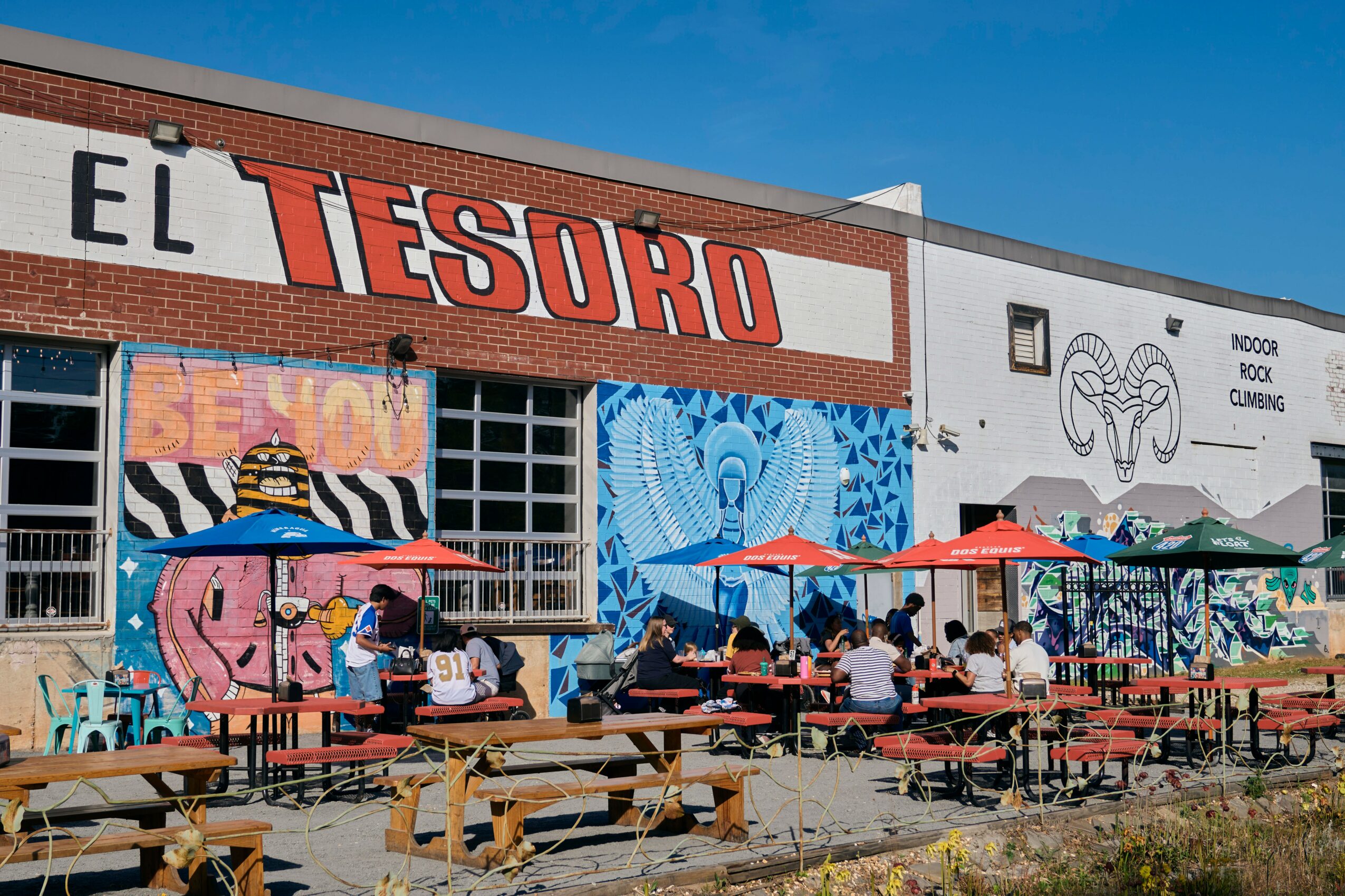 People sit at outdoor tables with umbrellas in front of a building with colorful murals and a sign reading 'EL TESORO'.