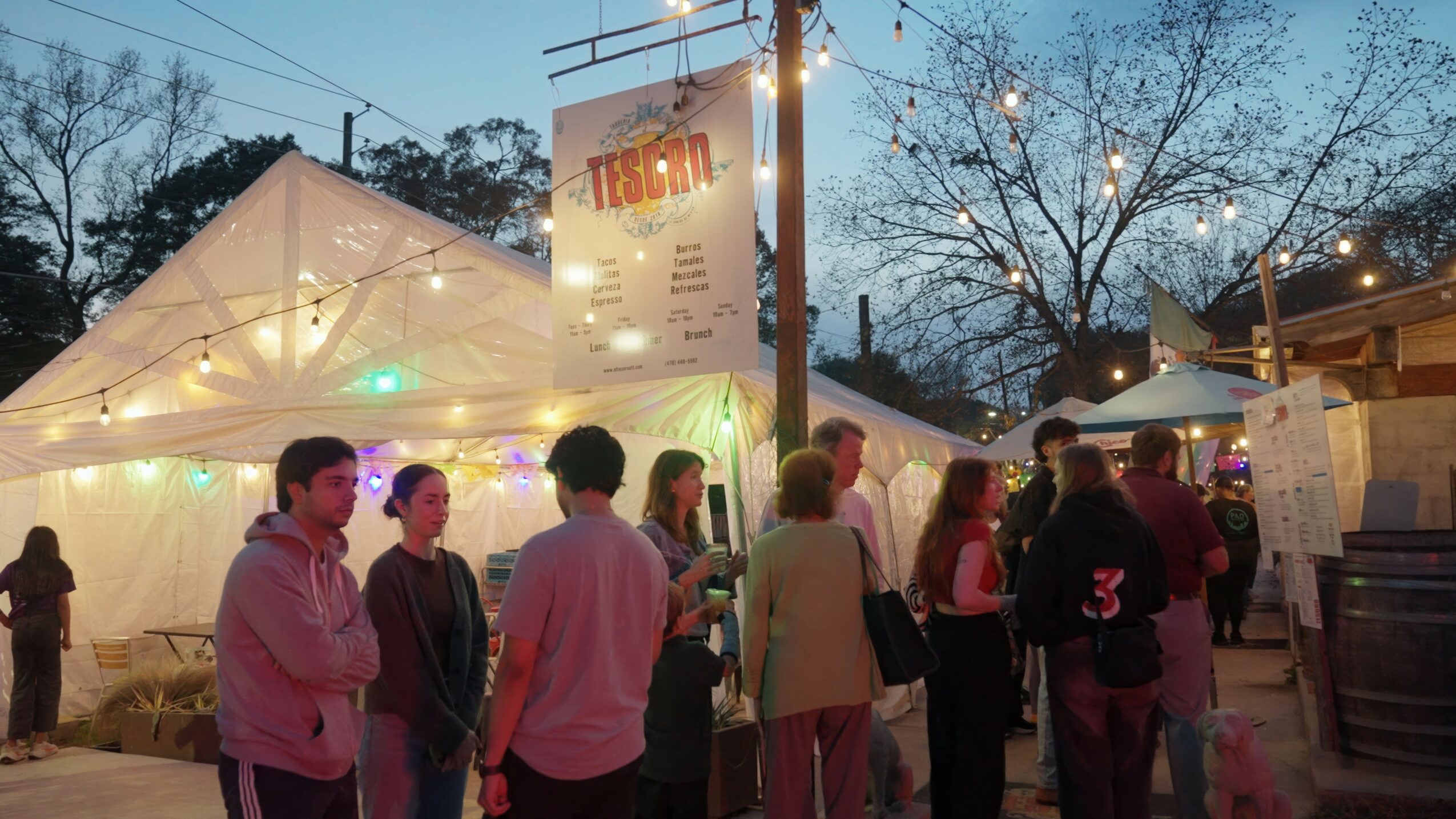 People gather outdoors near tents and a sign, with trees and evening sky in the background.