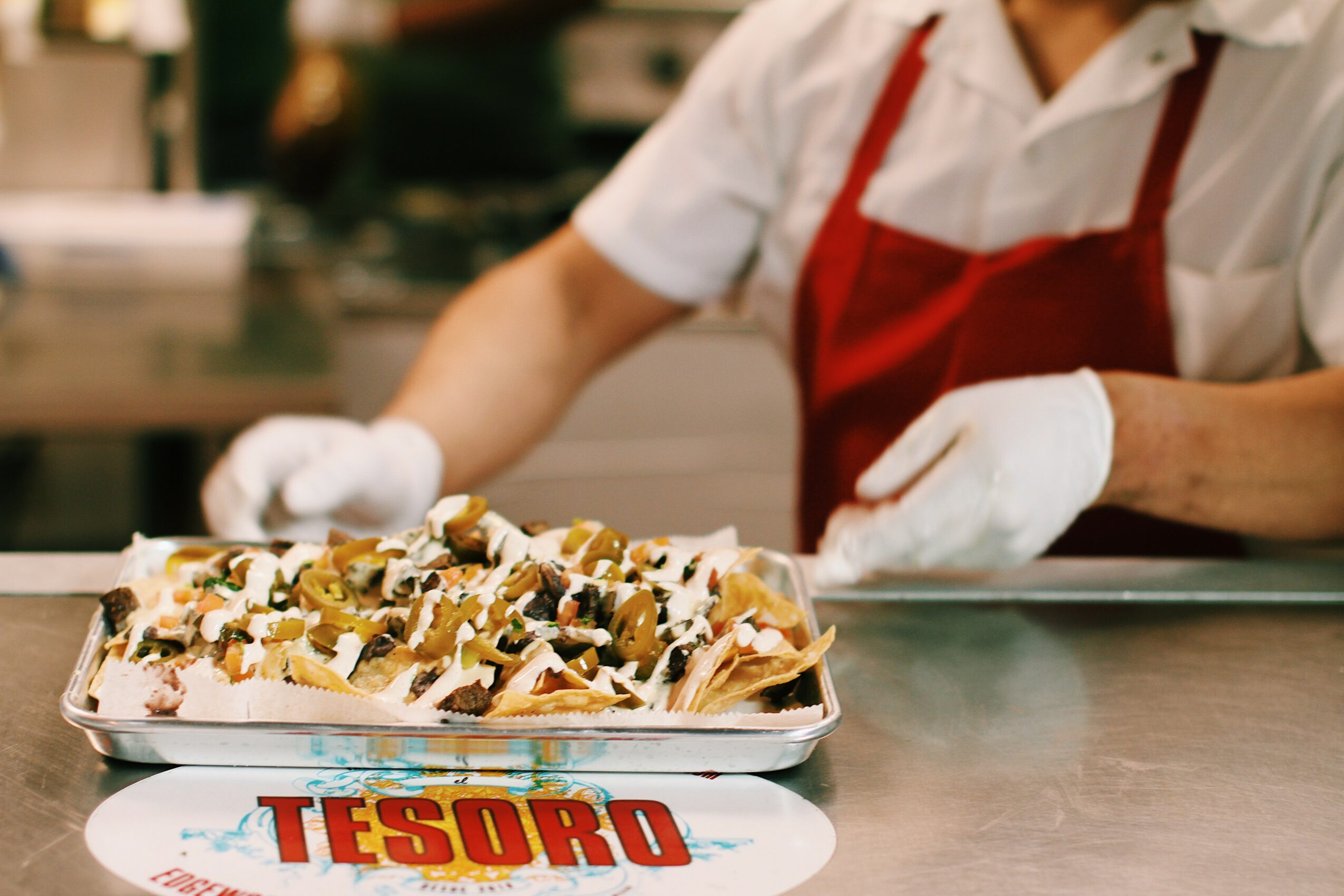 Person in white shirt and red apron preparing a tray of loaded fries with toppings on a stainless steel counter.