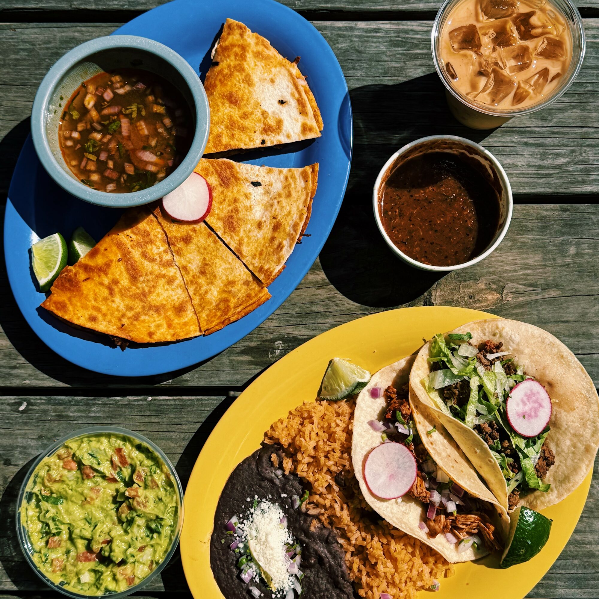 Plate with two tacos, guacamole, and rice, with drinks including iced coffee and a dark beverage, on a wooden table.