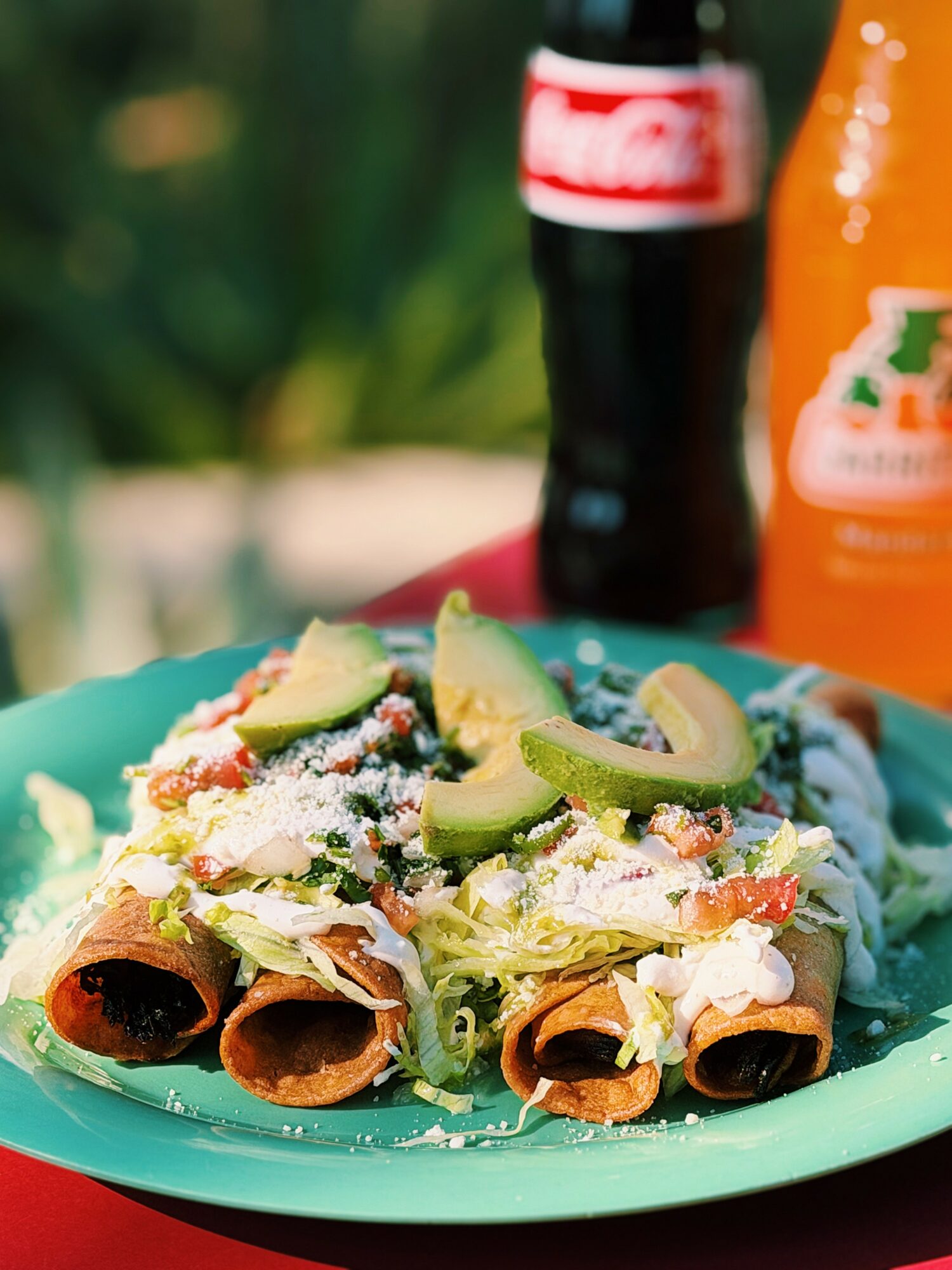 Plate with tacos topped with avocado slices, cheese, and lettuce, with a Coca-Cola bottle and orange drink in the background.