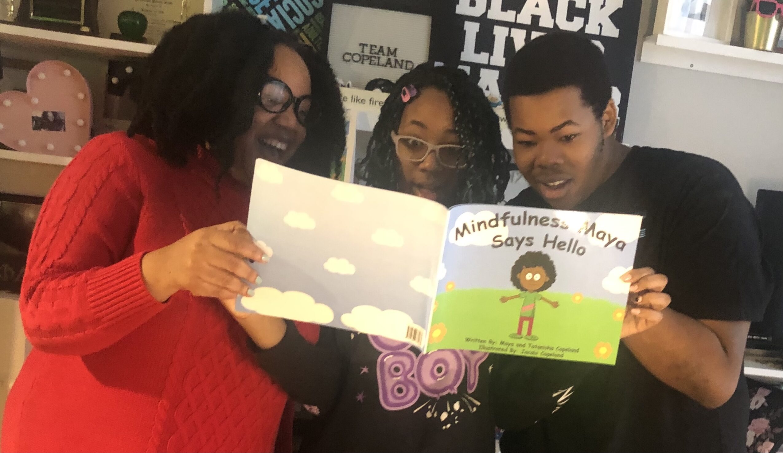 Three people reading a children's book together, smiling, in a room with posters and shelves.