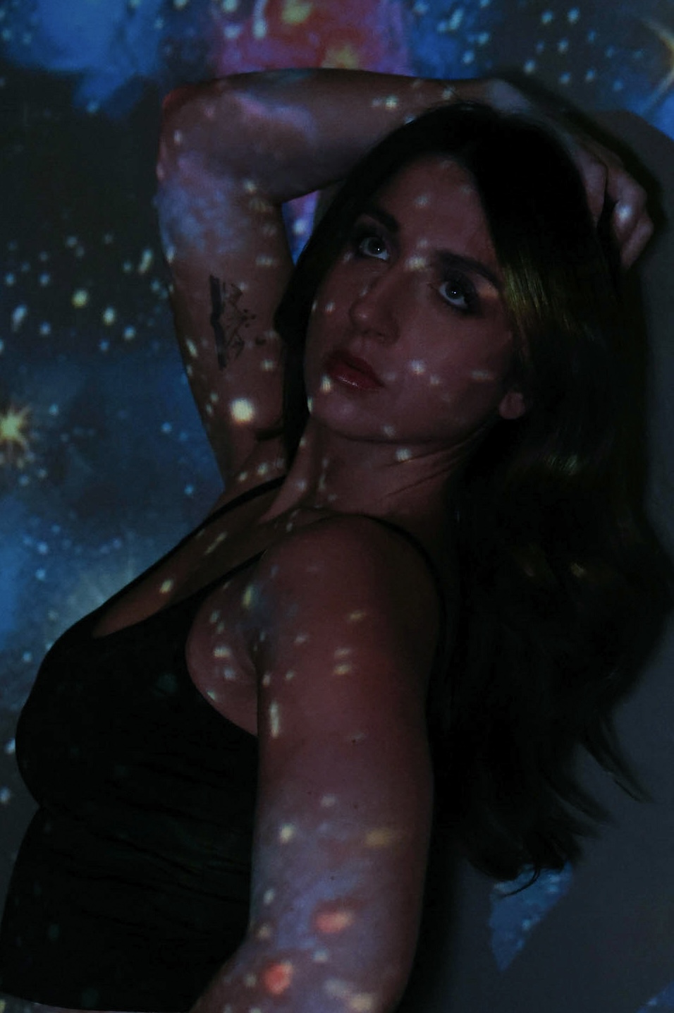 Woman with long dark hair poses with arm behind head, illuminated by colorful light projections, looking at camera.