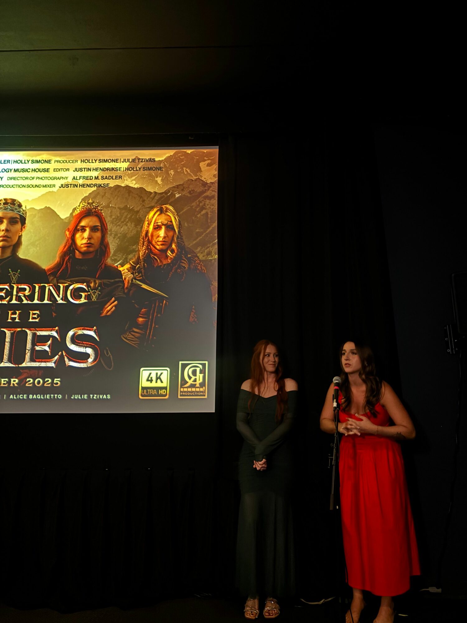 Three people stand in front of a black curtain, with a poster for 'The Mays' behind them. One woman wears a red dress.