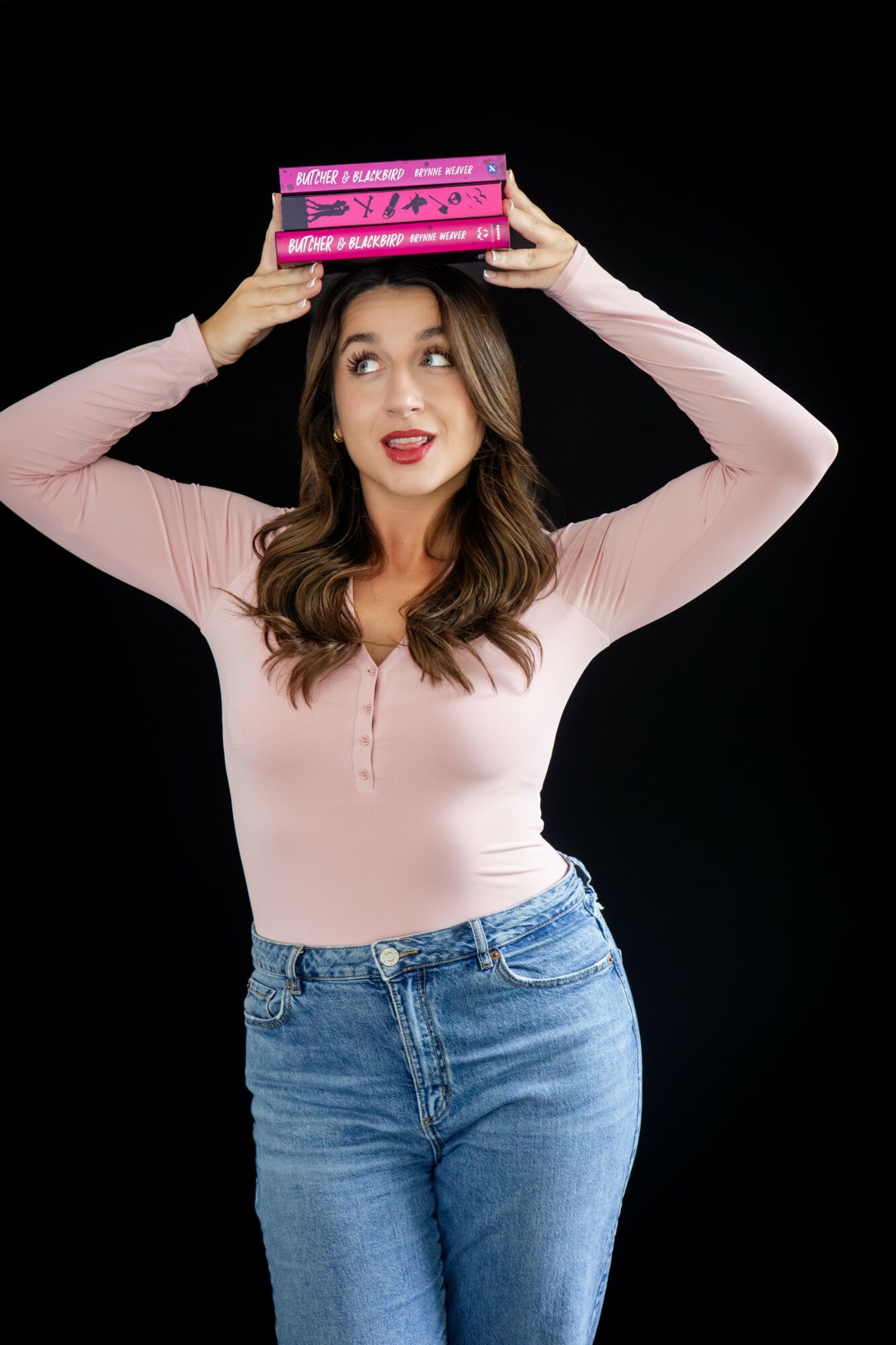 Young woman with long brown hair balancing pink books on her head, wearing a light pink top and blue jeans, against a black background.