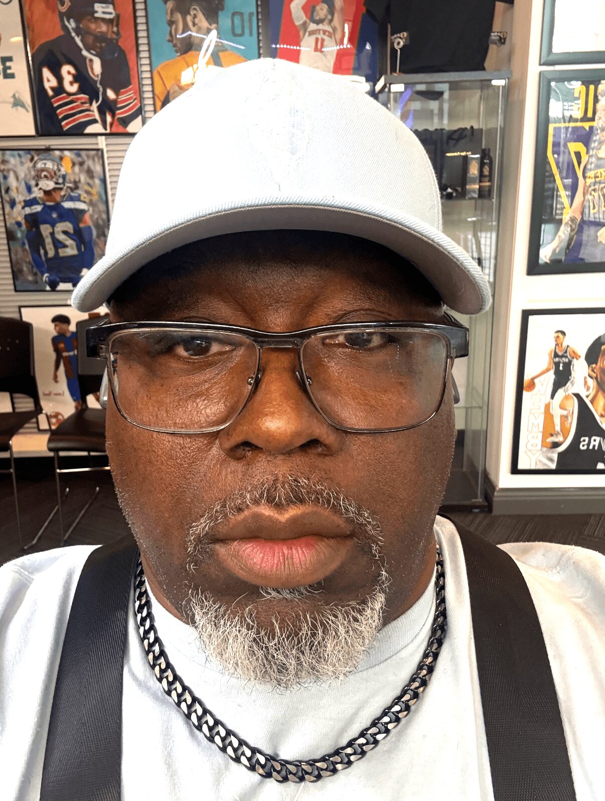 Man wearing glasses, white cap, and a chain necklace, with sports posters in the background.