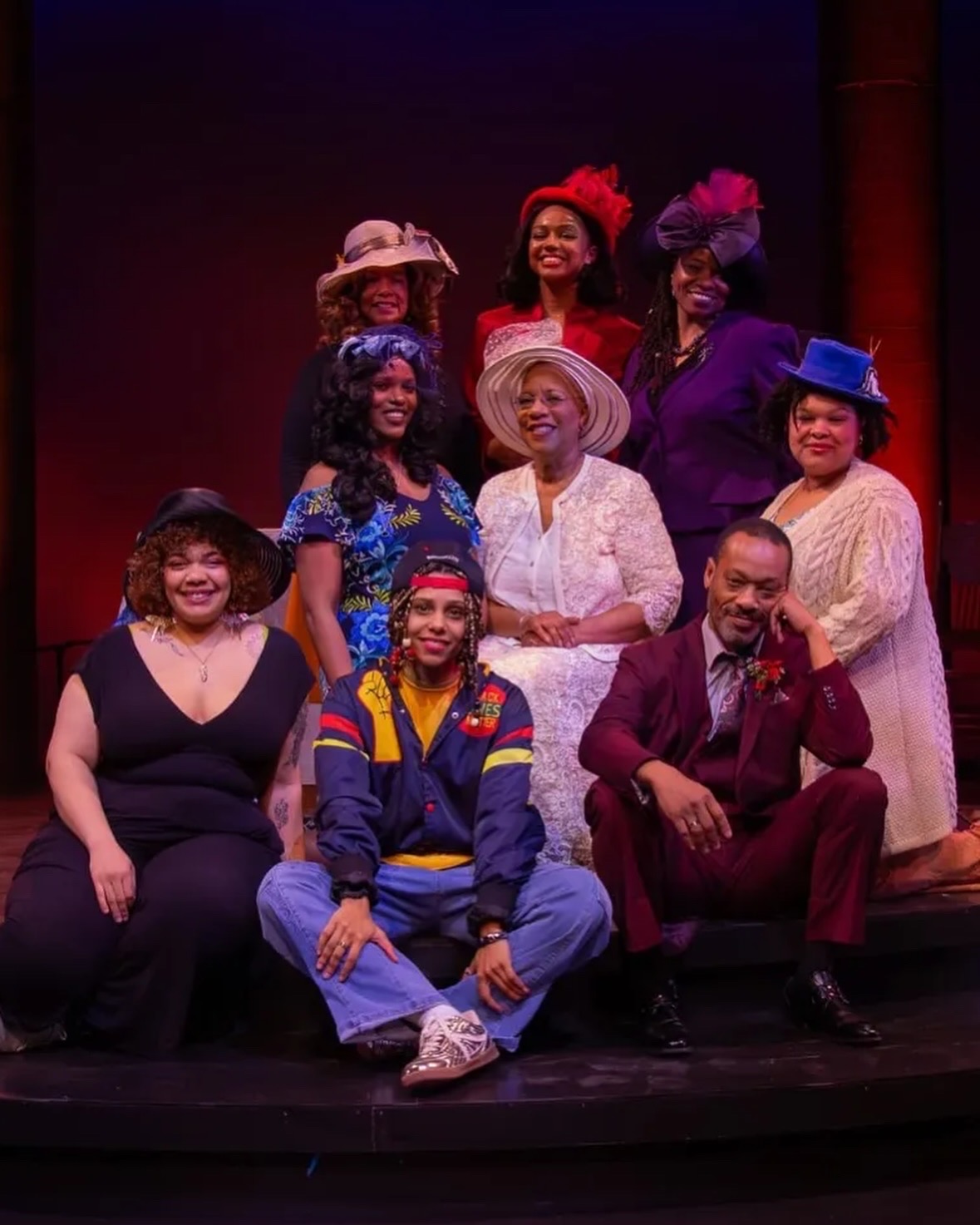 Group of nine people on stage, dressed in colorful costumes and hats, smiling and posing for a photo.