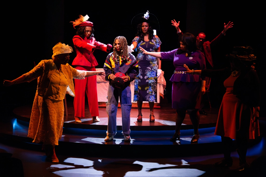 Group of people on stage performing, some with arms raised, dressed in colorful costumes, with a dark background.