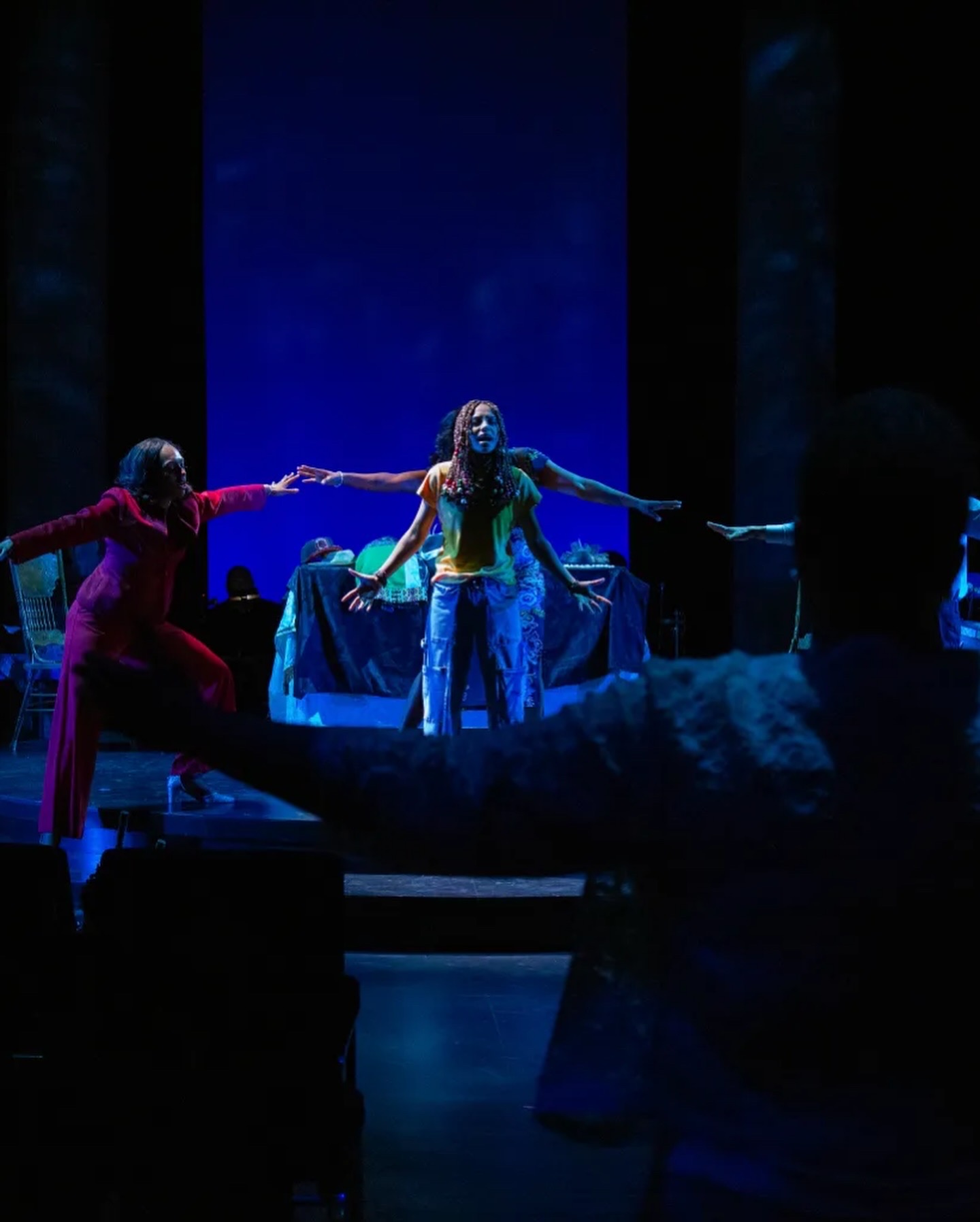 Three performers on stage holding hands in a dance or theatrical performance, with a blue background and audience in foreground.