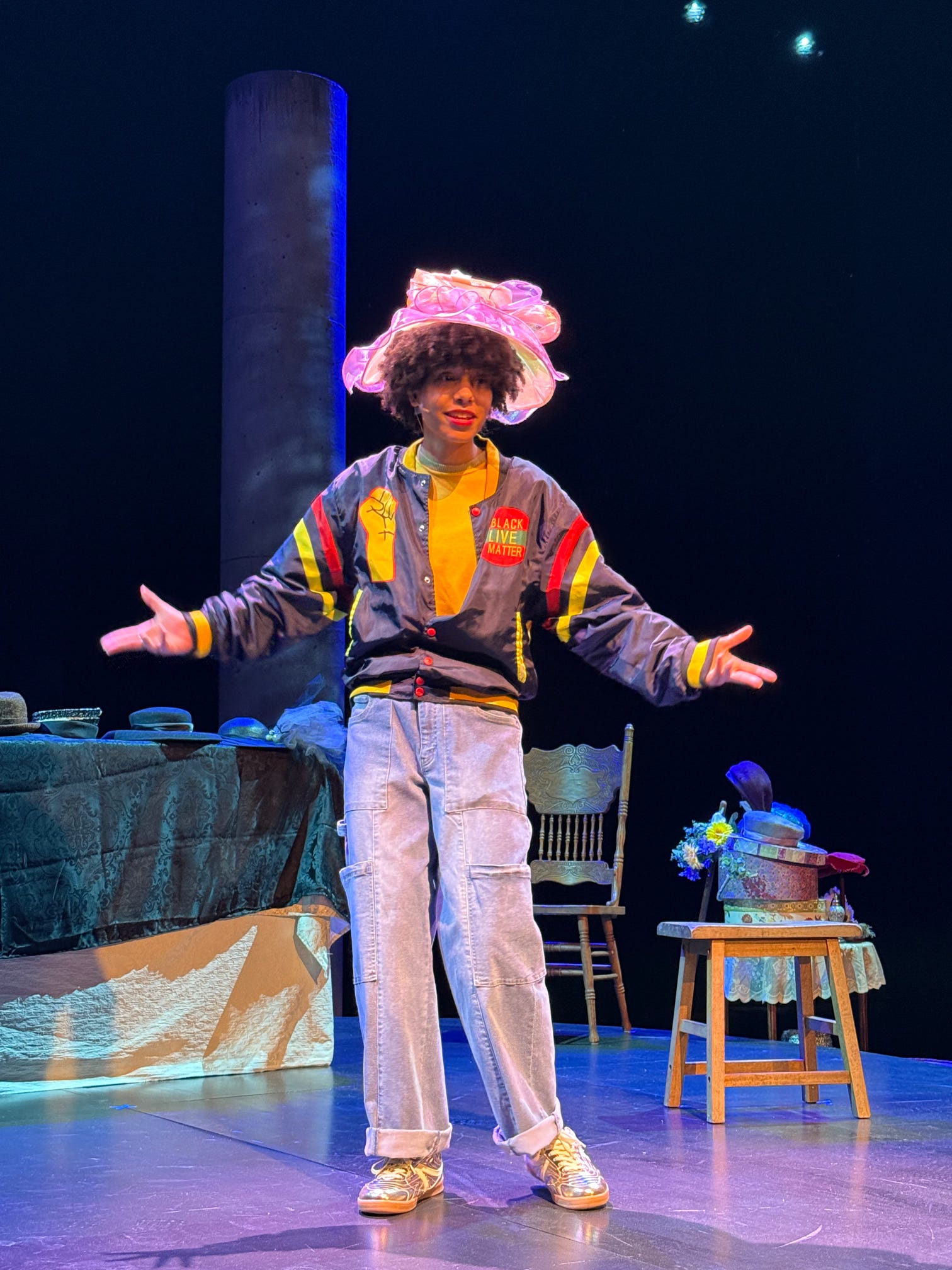 Person with curly hair wearing a colorful jacket, light jeans, and a pink hat, standing on stage with arms outstretched.