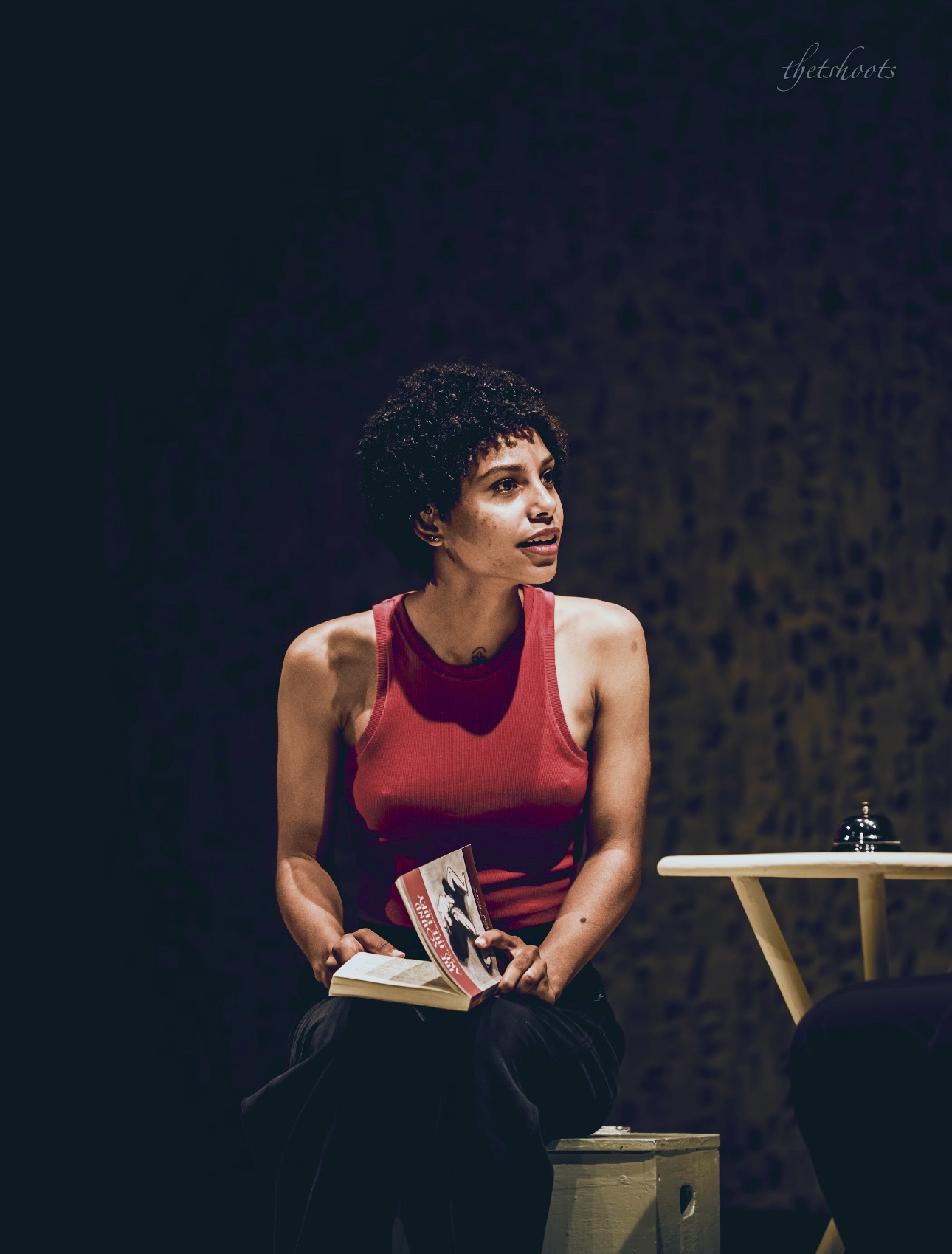 Woman with short curly hair wearing a red sleeveless top, sitting on a stool, holding a book, against a dark background.
