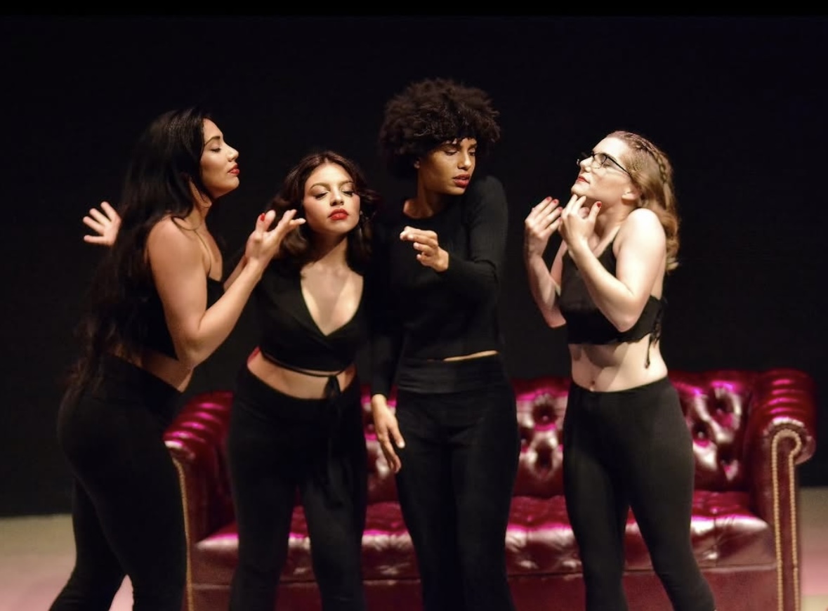 Four women stand on stage with a pink velvet sofa behind them, engaged in conversation or performance, against a dark background.