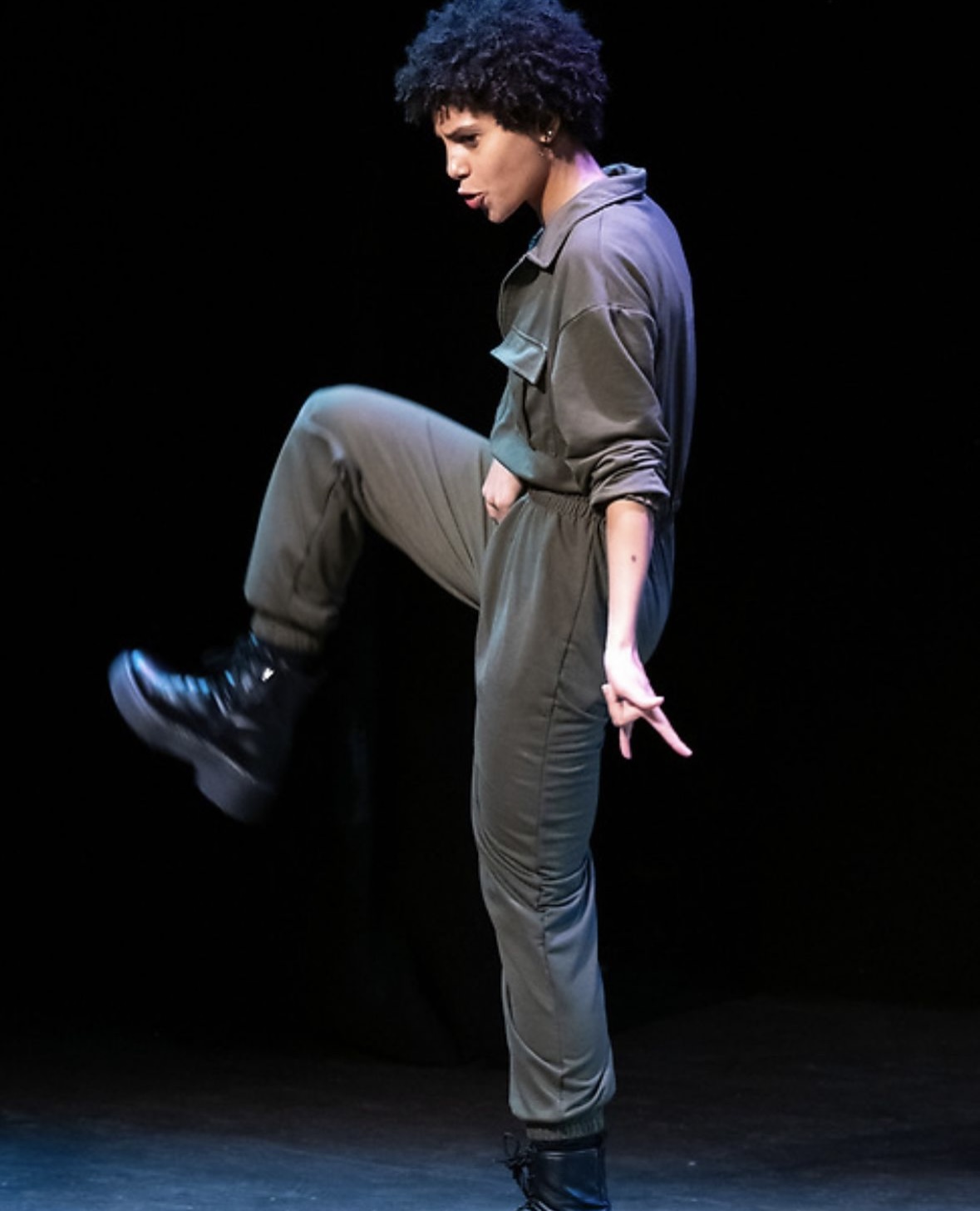 Person with curly hair in gray jumpsuit balancing on one leg, dark background, performing dance move.