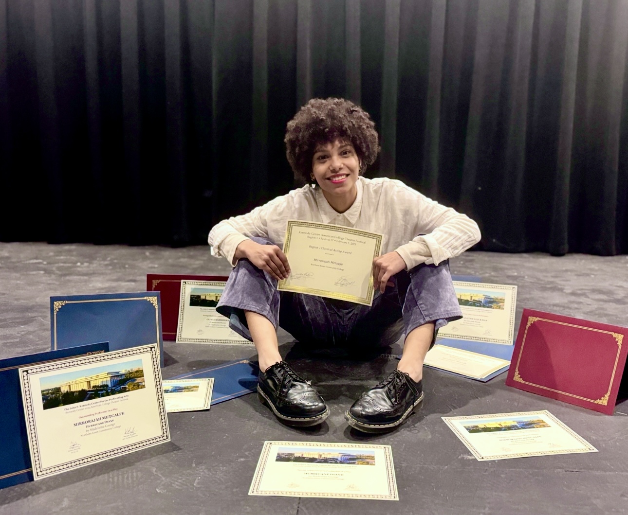 Person sitting on stage holding a certificate, surrounded by other certificates, with a black curtain background.