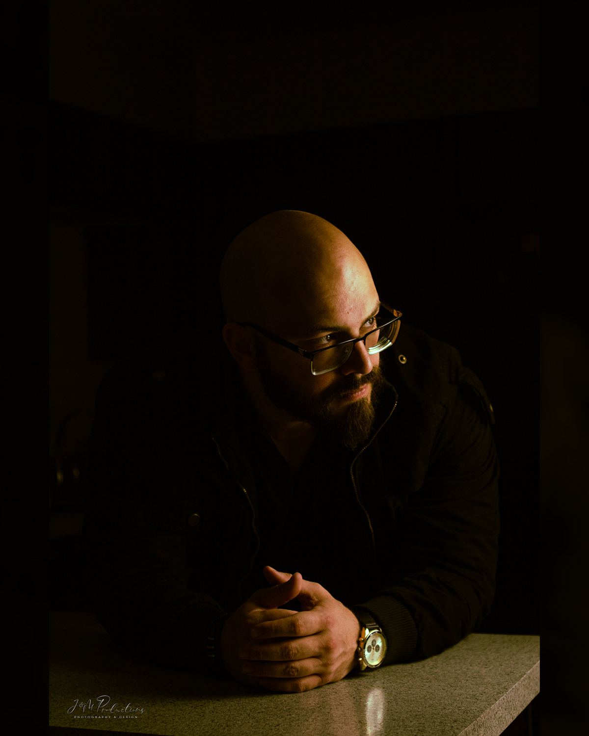 Bald man with glasses and beard sitting at a table, hands clasped, in a dark setting.
