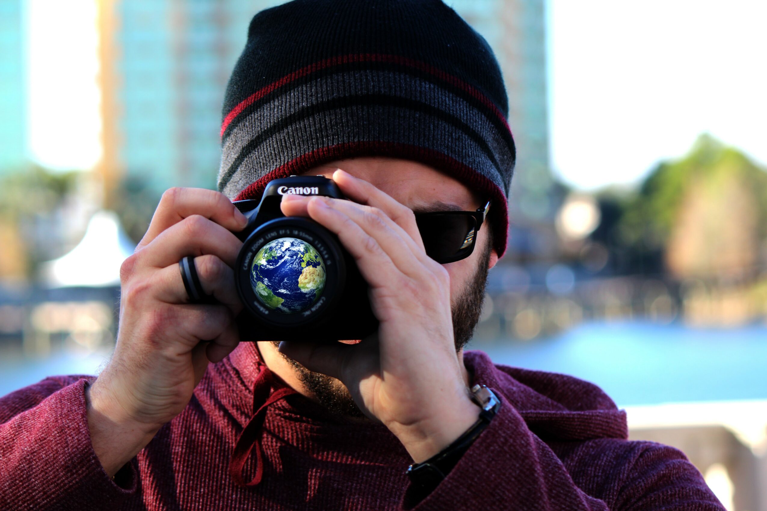 Person wearing a beanie and sunglasses taking a photo with a camera outdoors, with water and buildings in the background.