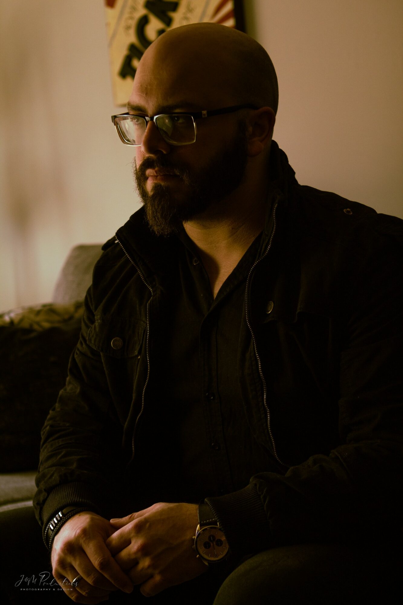 Man with glasses and a beard, wearing a dark jacket, sitting indoors with warm lighting.