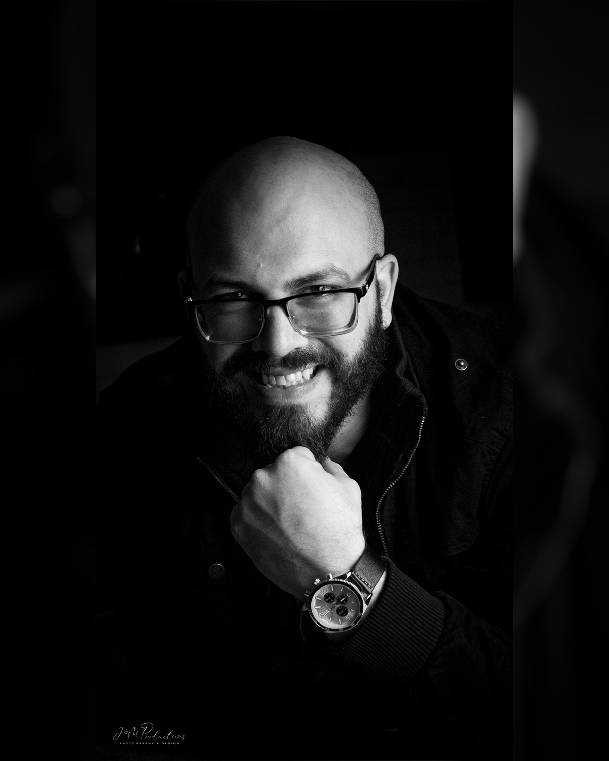 Bald man with glasses smiling, resting chin on hand, wearing a watch and dark clothing, in black and white.