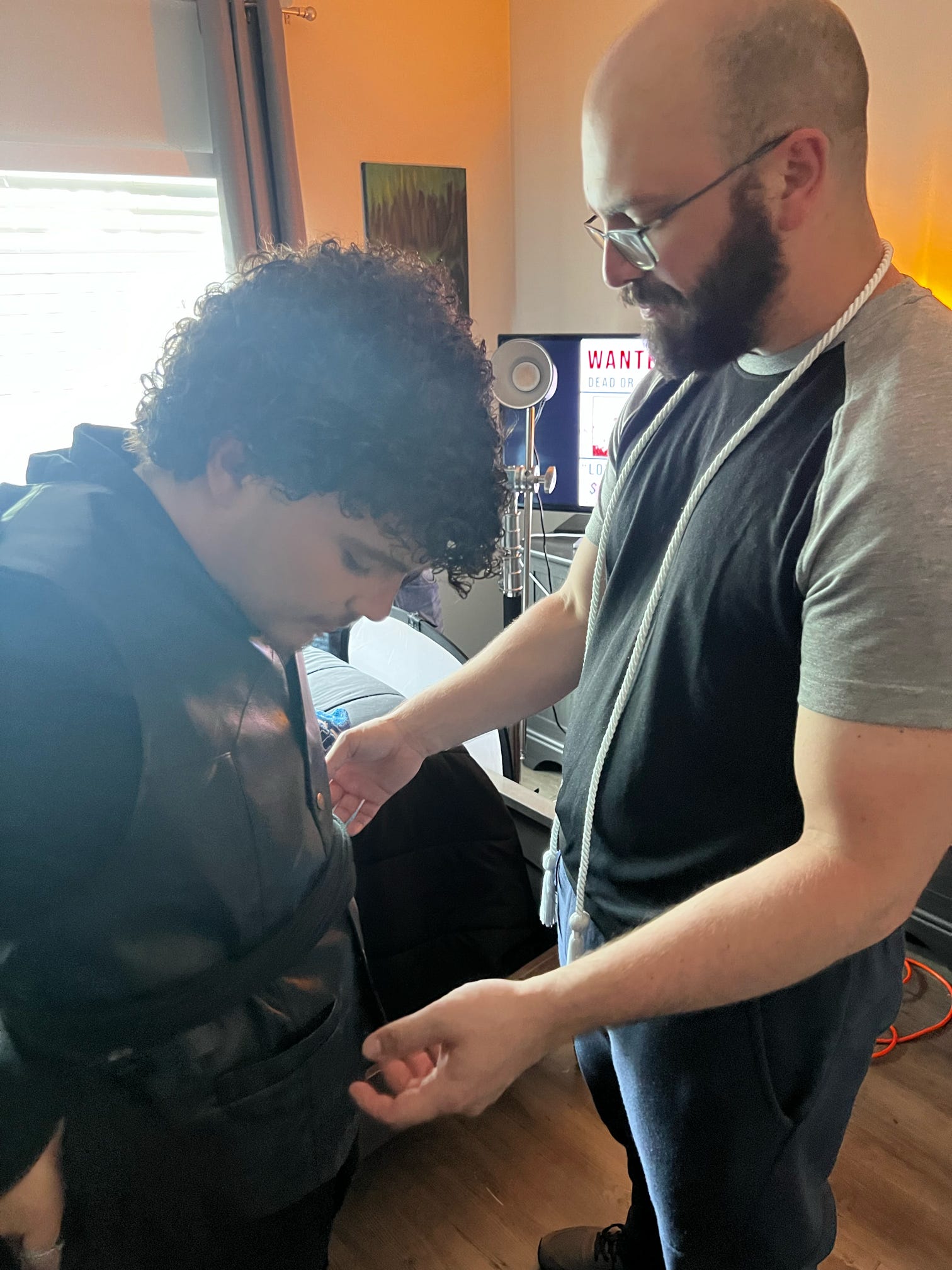 Two people standing close, one with curly hair and the other with glasses and a beard, in a room with electronic equipment.