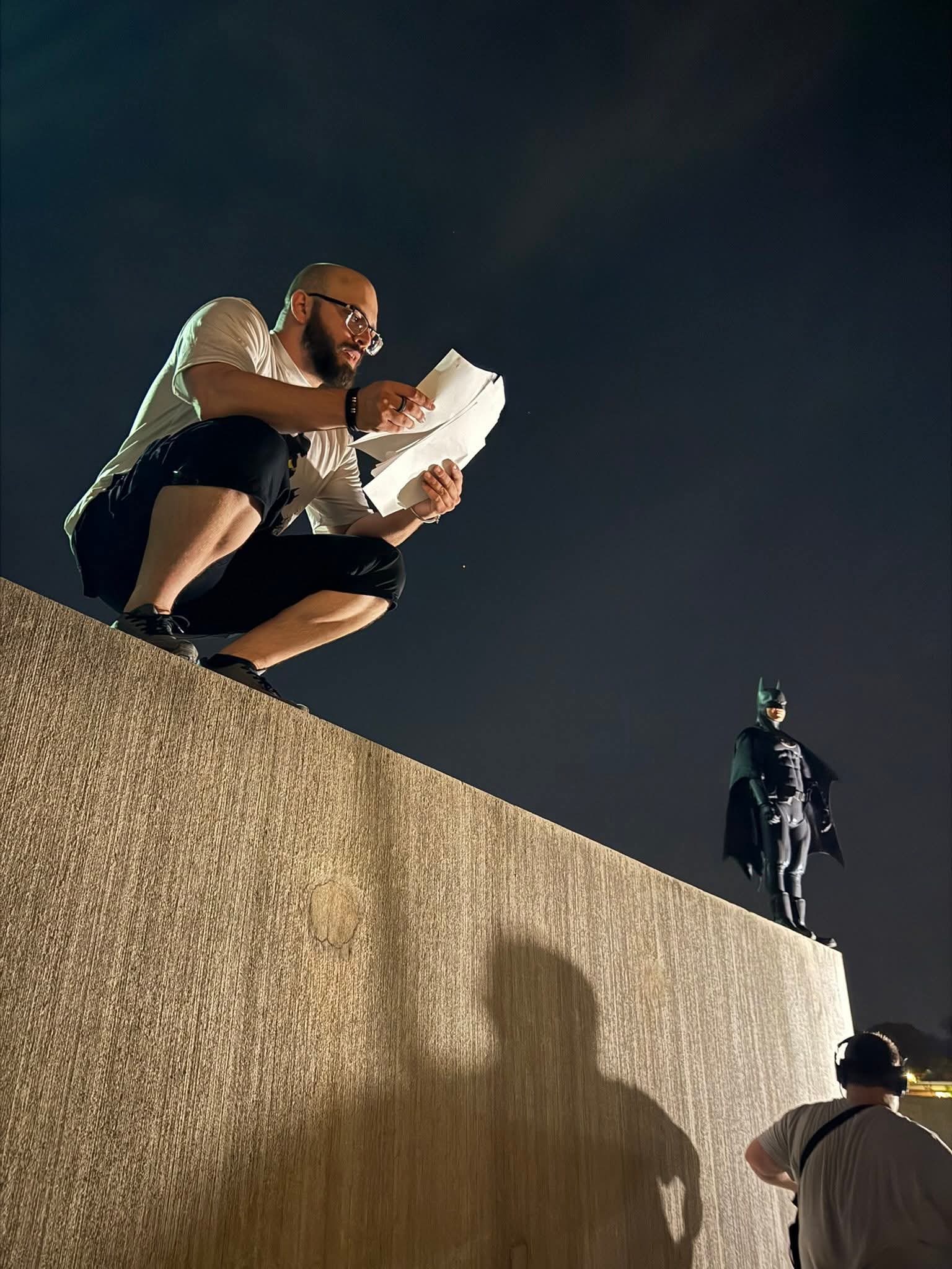 Man crouching on a ledge reading a book, with a statue and another person in the background at night.