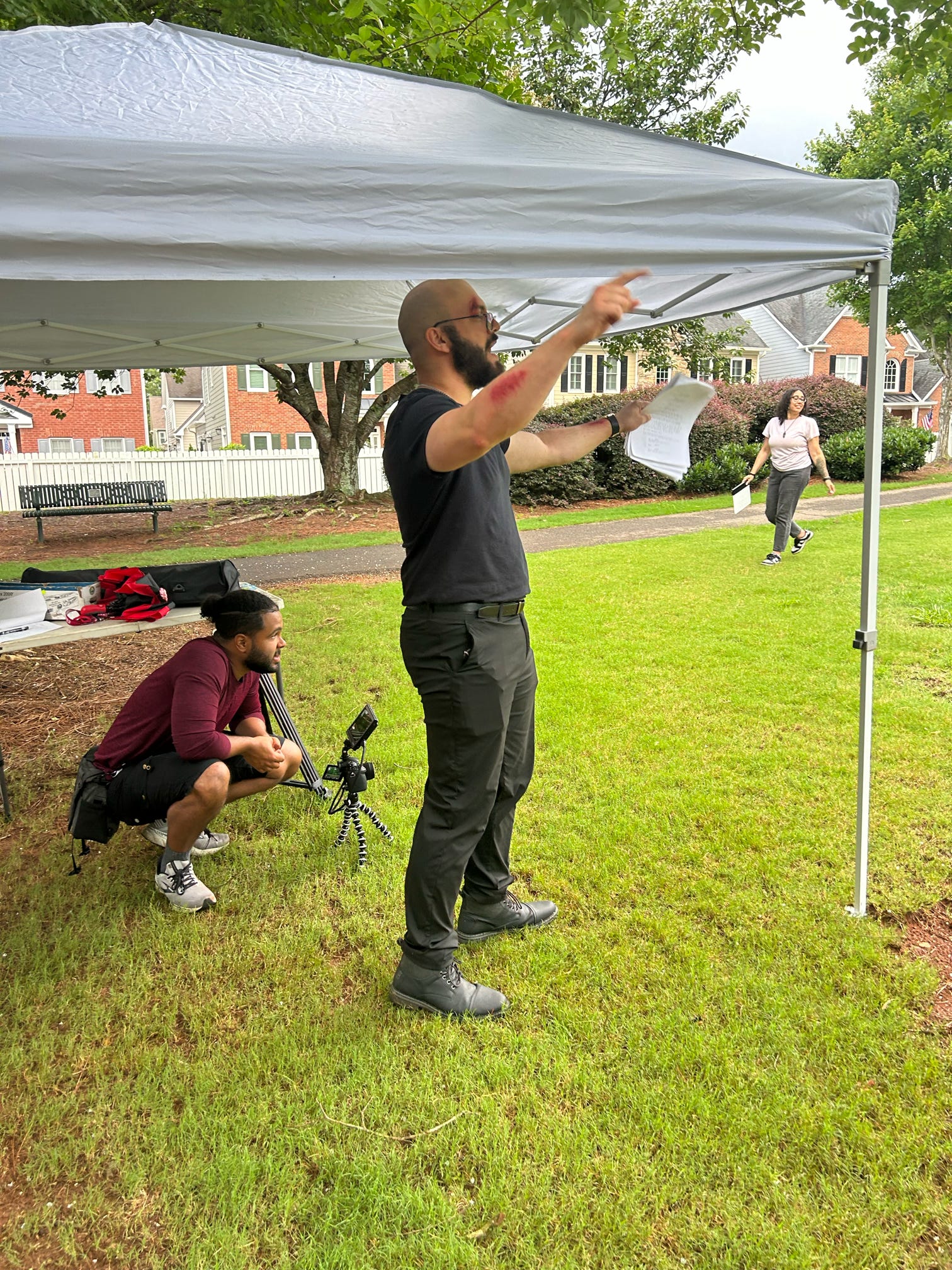 Man standing under a canopy, gesturing with hands, holding papers, in a park with grass and trees.