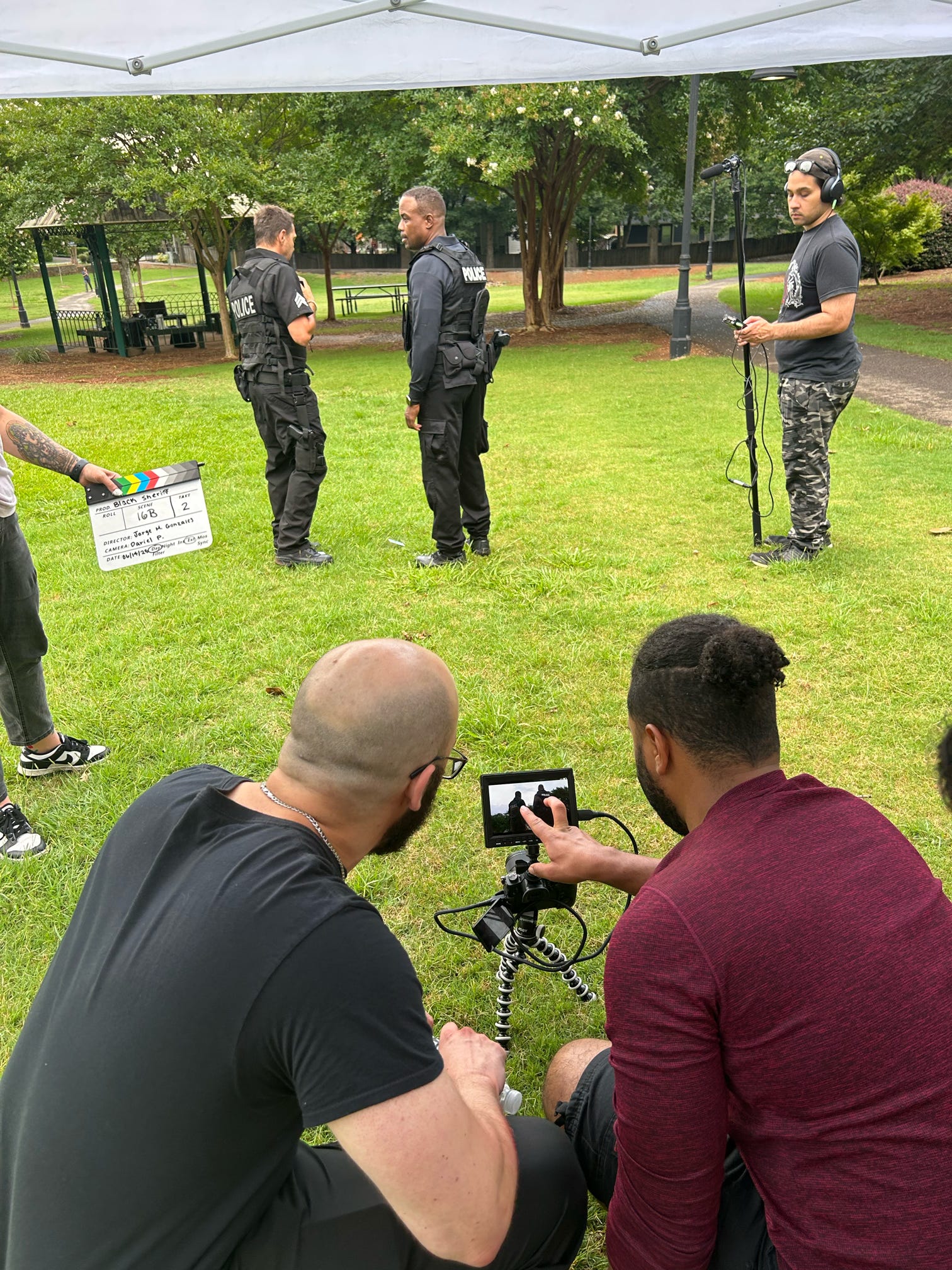 Three people in black uniforms stand on grass under a canopy, with two men filming them from the foreground, trees in background.