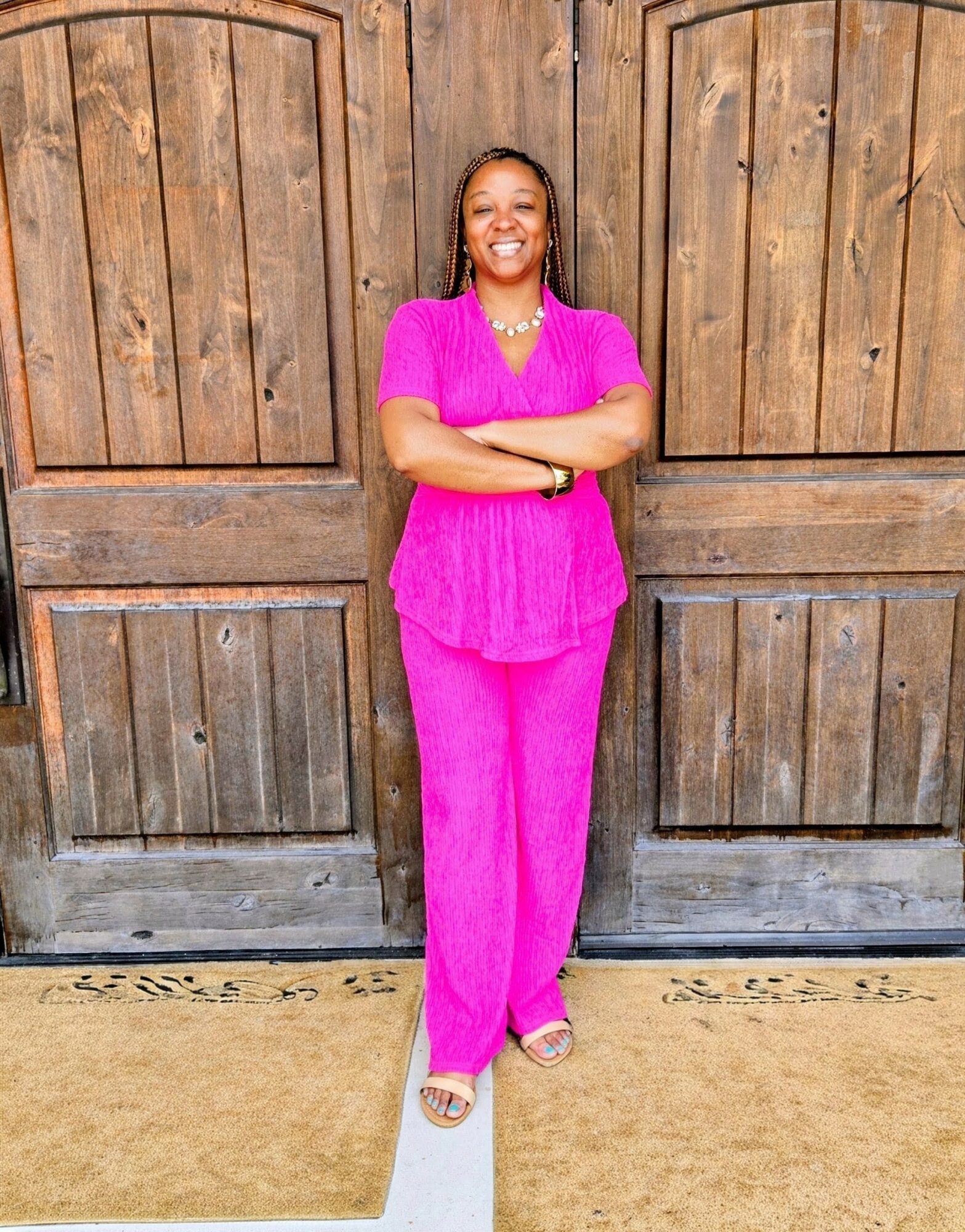 Smiling woman in pink outfit standing with arms crossed in front of wooden doors, smiling, on a beige and white floor.