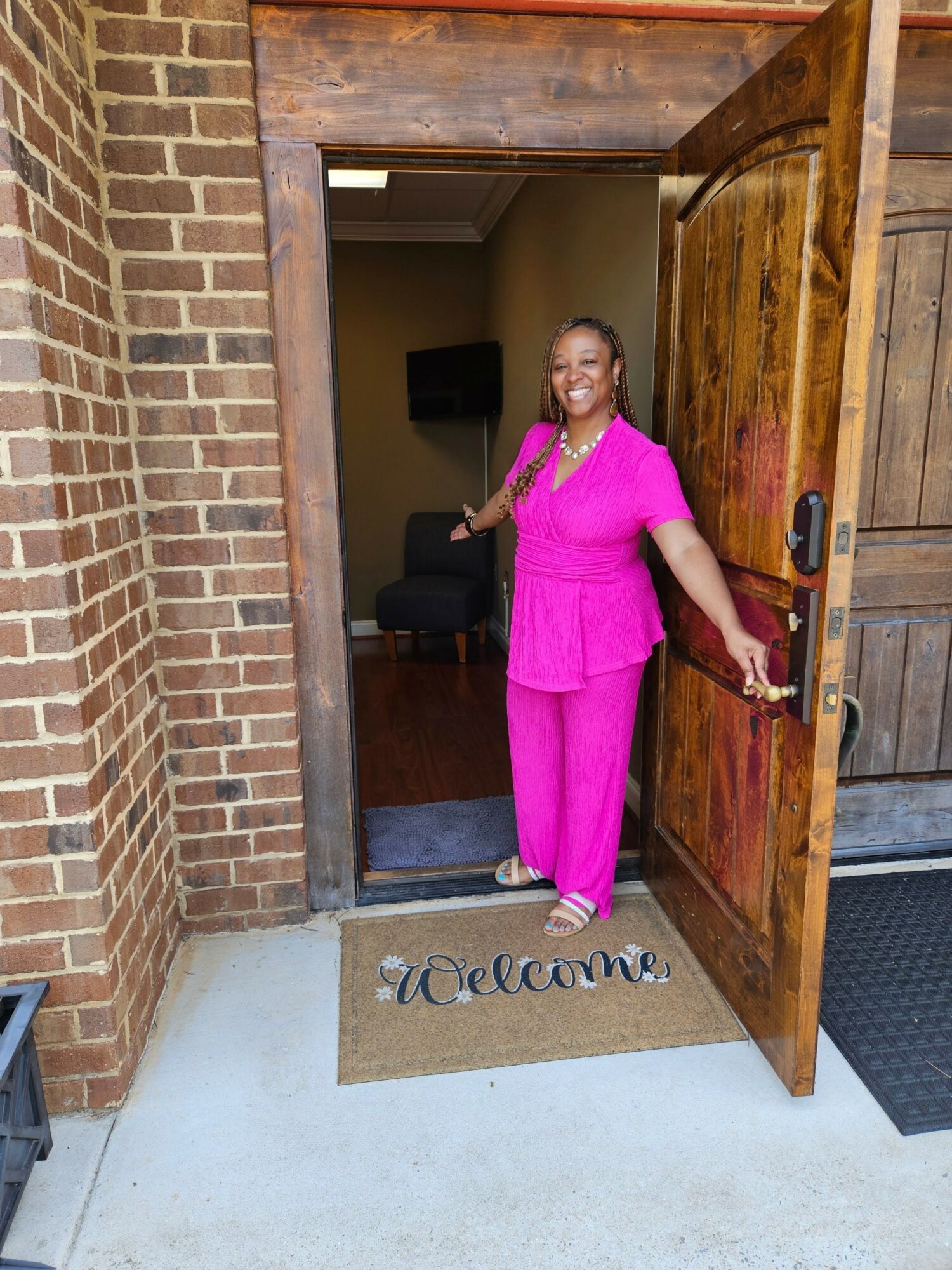 Woman in pink outfit standing at open wooden door, smiling, inside a brick building, welcome mat on floor.