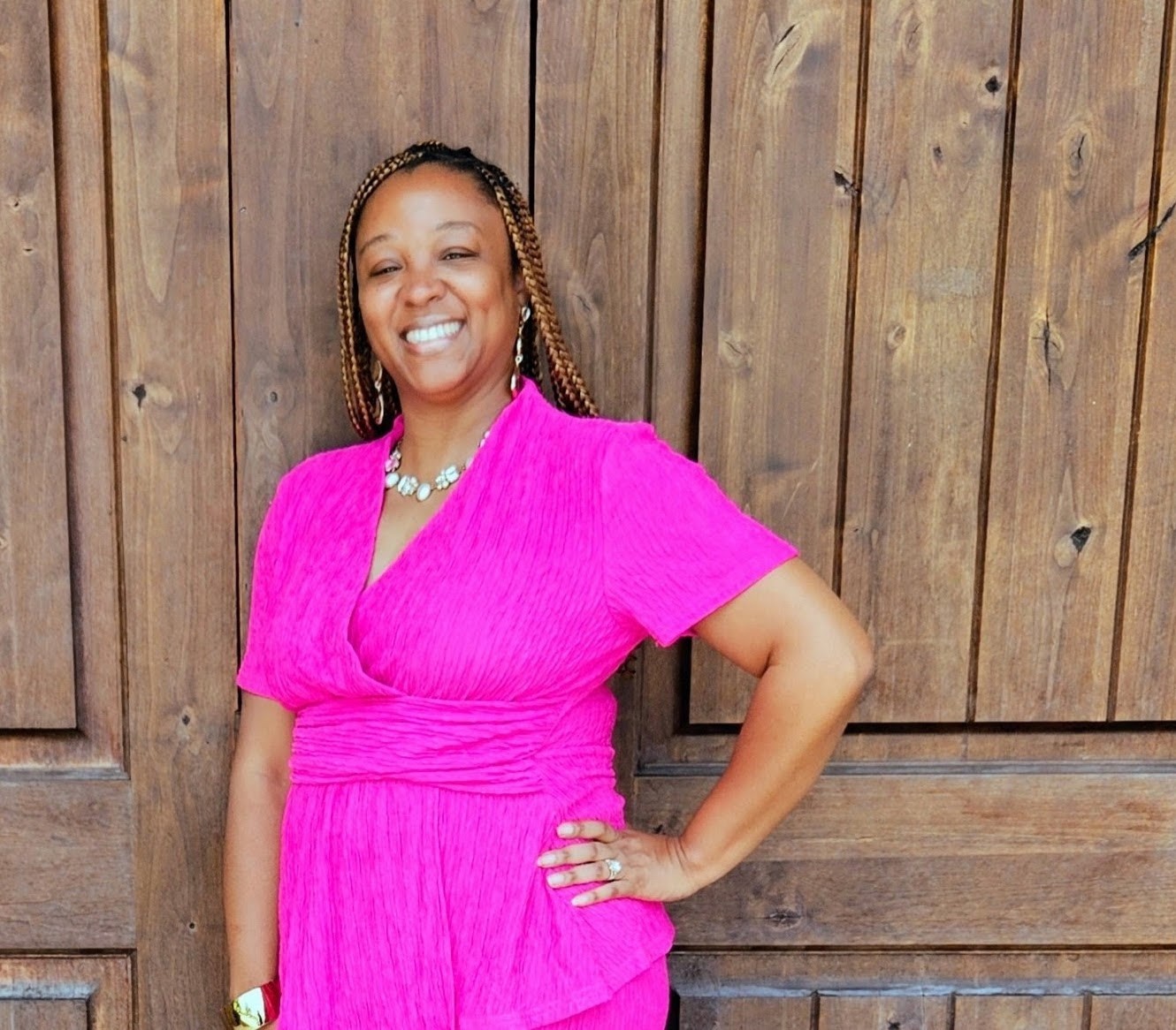 Woman in pink dress smiling with hand on hip in front of wooden fence.