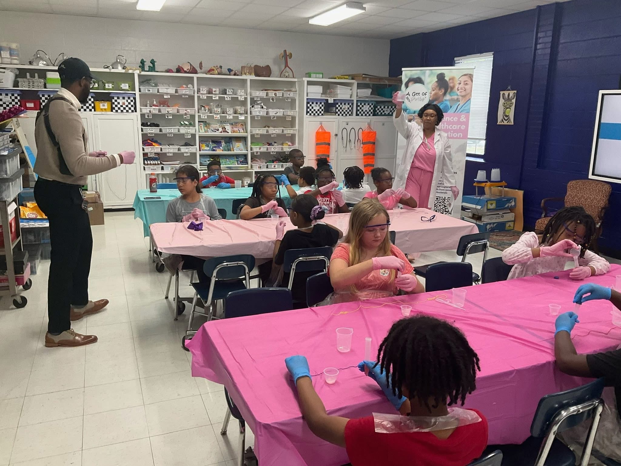 Children seated at pink-covered tables in a classroom, with a teacher and an adult standing nearby, shelves with supplies in the background.