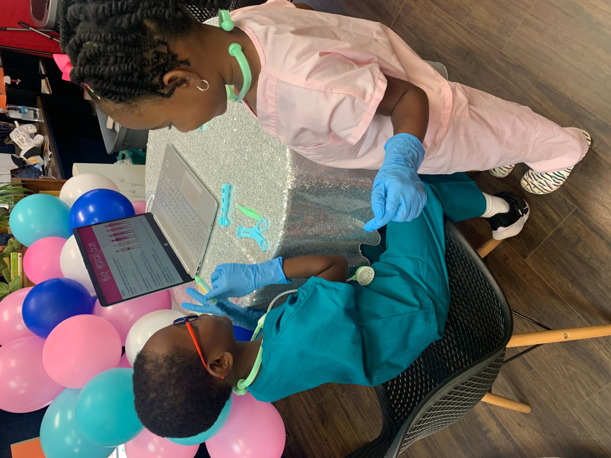 Two children in protective clothing work on a cake decorated with balloons, using a laptop and tools, on a table.