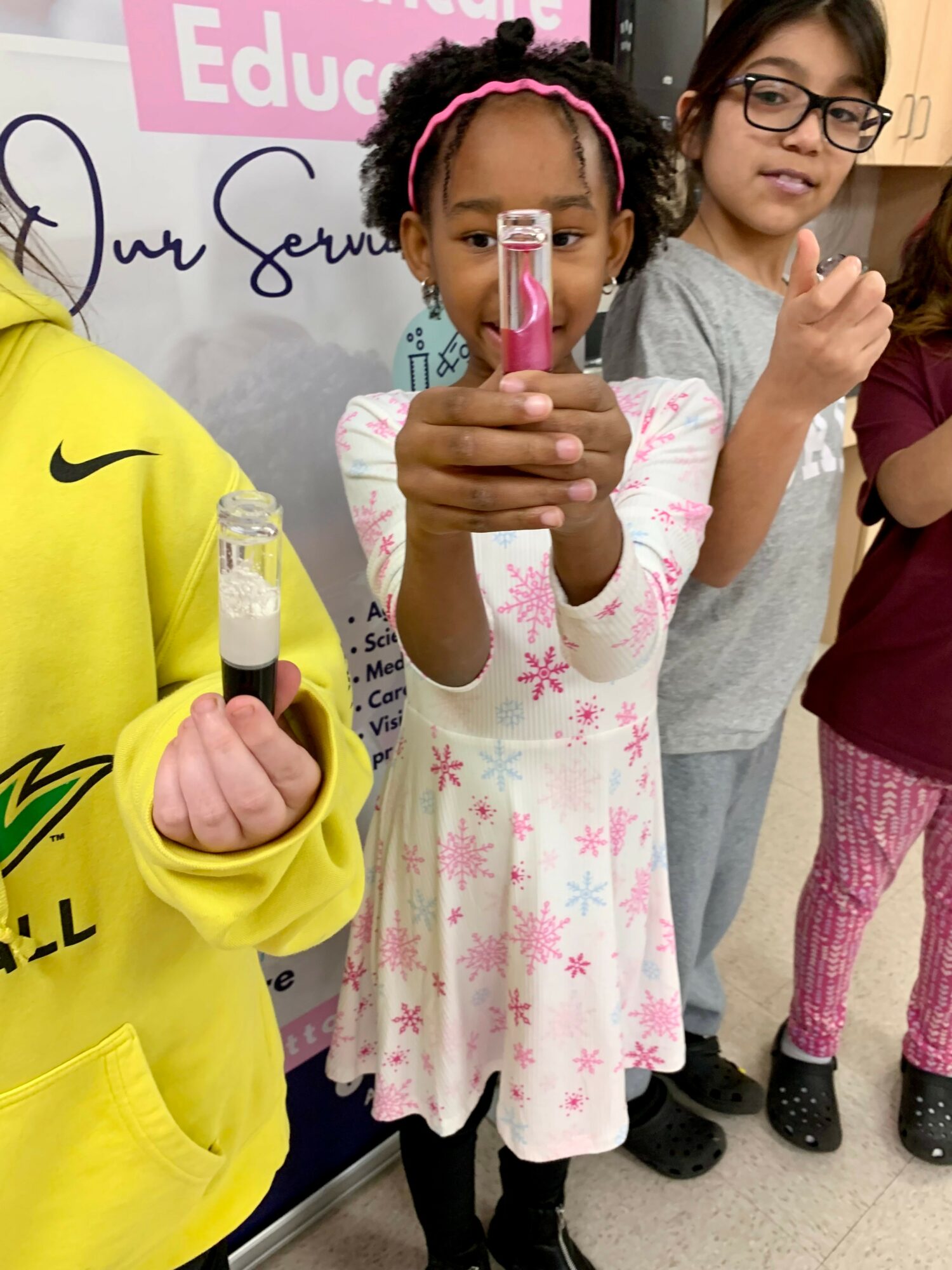 Child holding a pink object, with two other children nearby, in a classroom setting.