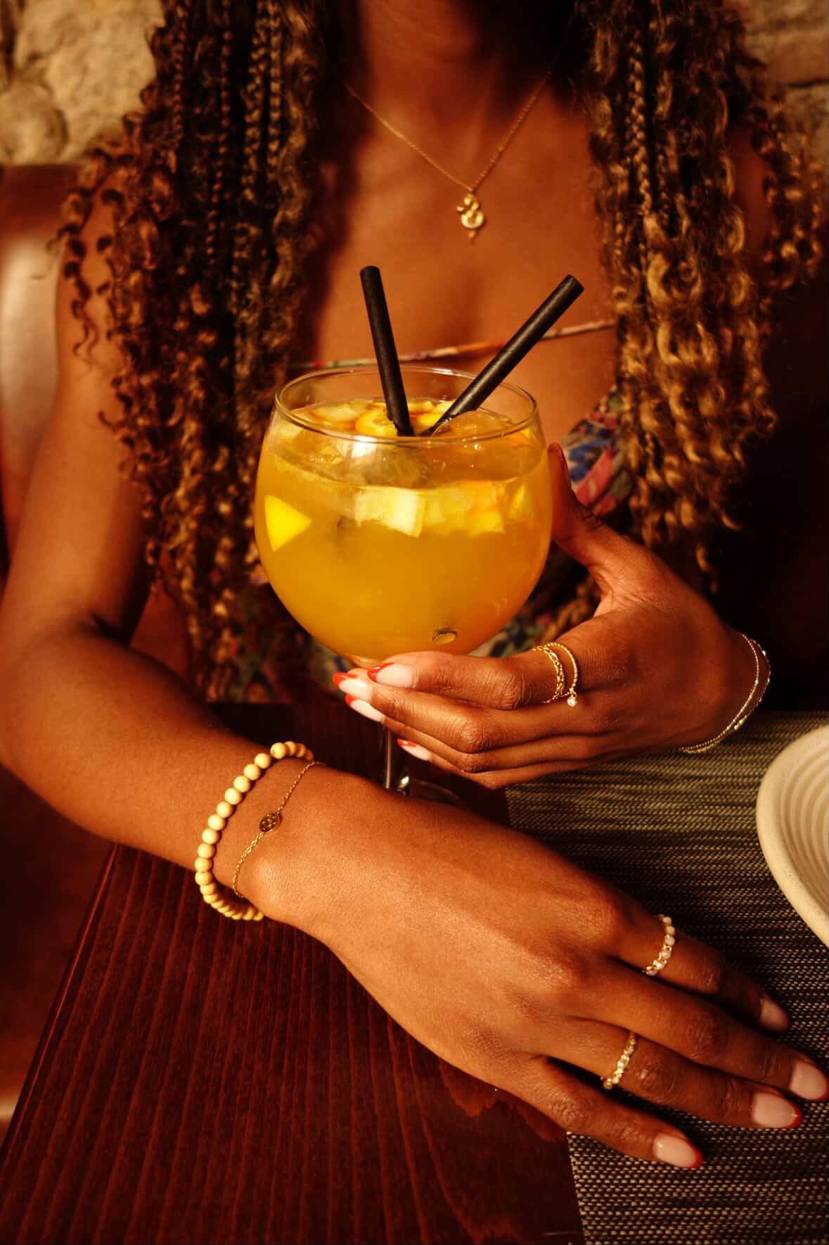 Person holding a large yellow cocktail with ice and two black straws, wearing jewelry, with curly hair and a dark top.