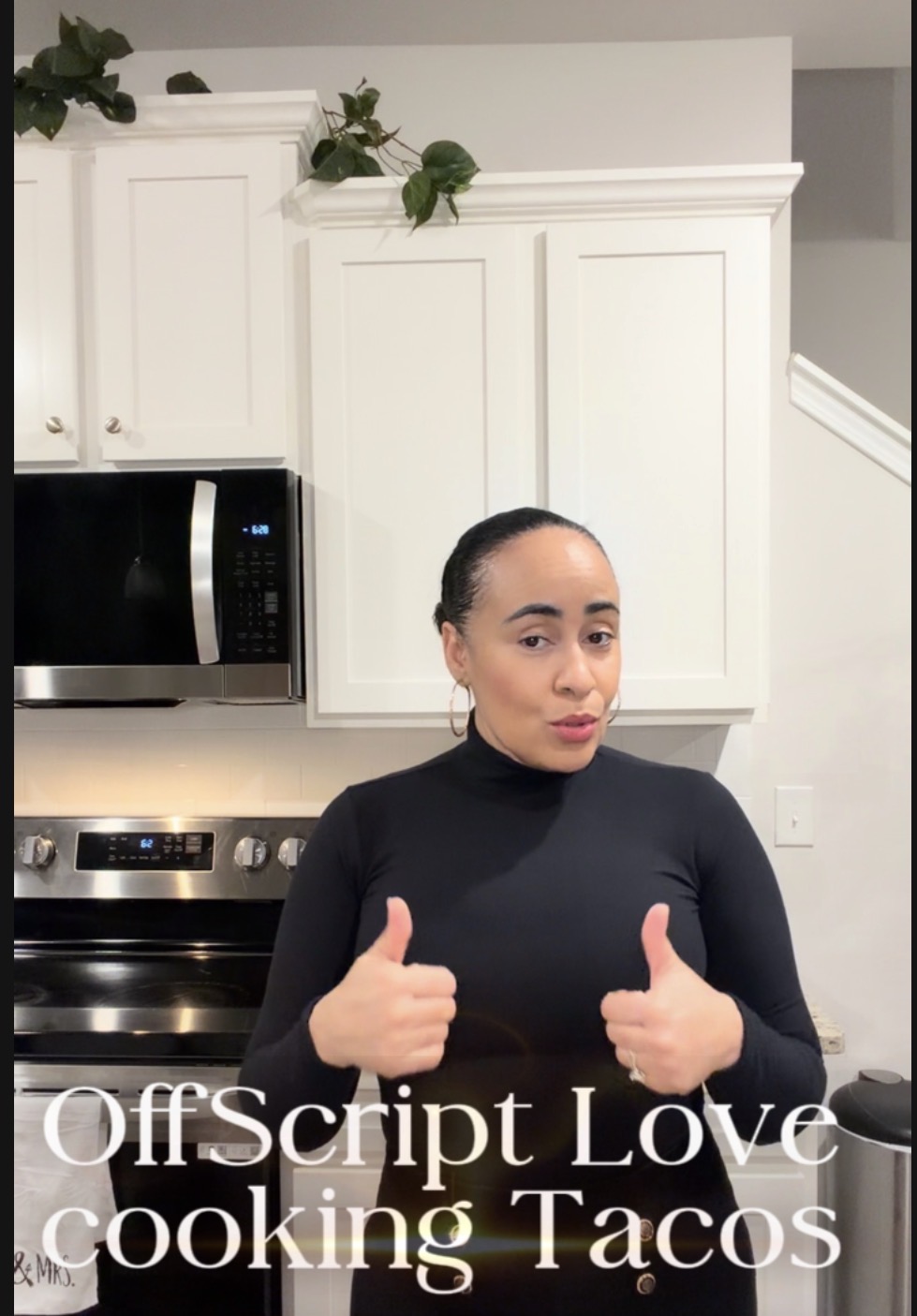 Woman in black turtleneck giving two thumbs up in a kitchen with white cabinets and stainless steel appliances.