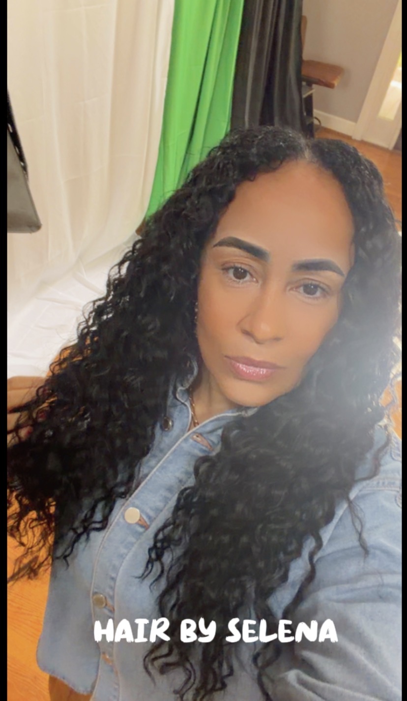 Close-up of a woman with long, curly hair wearing a denim jacket, indoors with colorful curtains in the background.