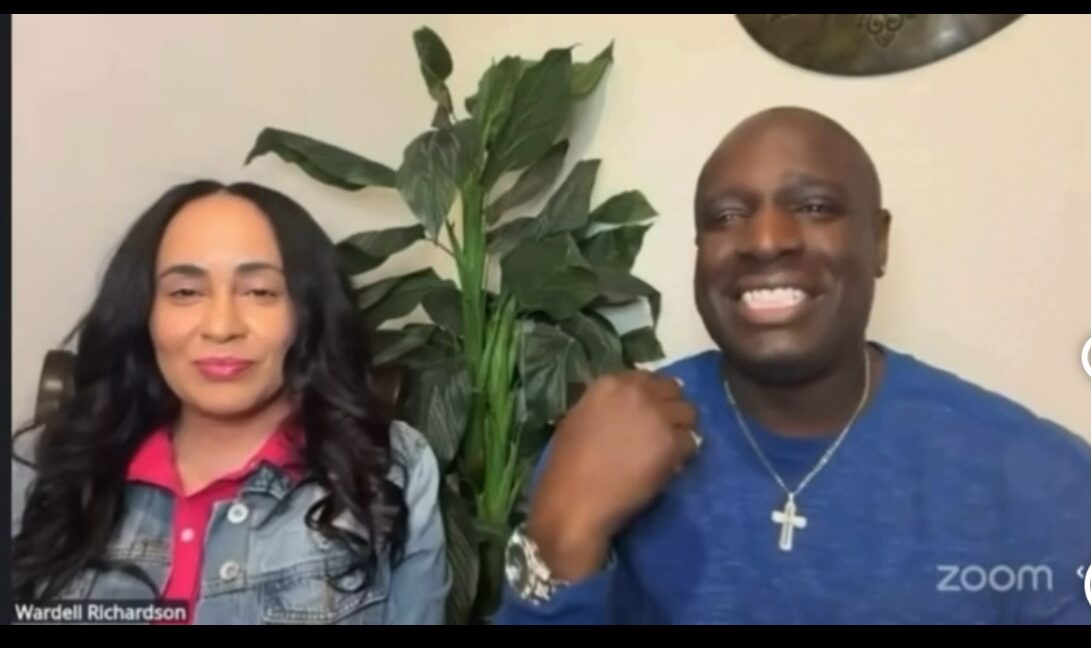 Two people sitting in front of a plant, smiling, with a white wall background.