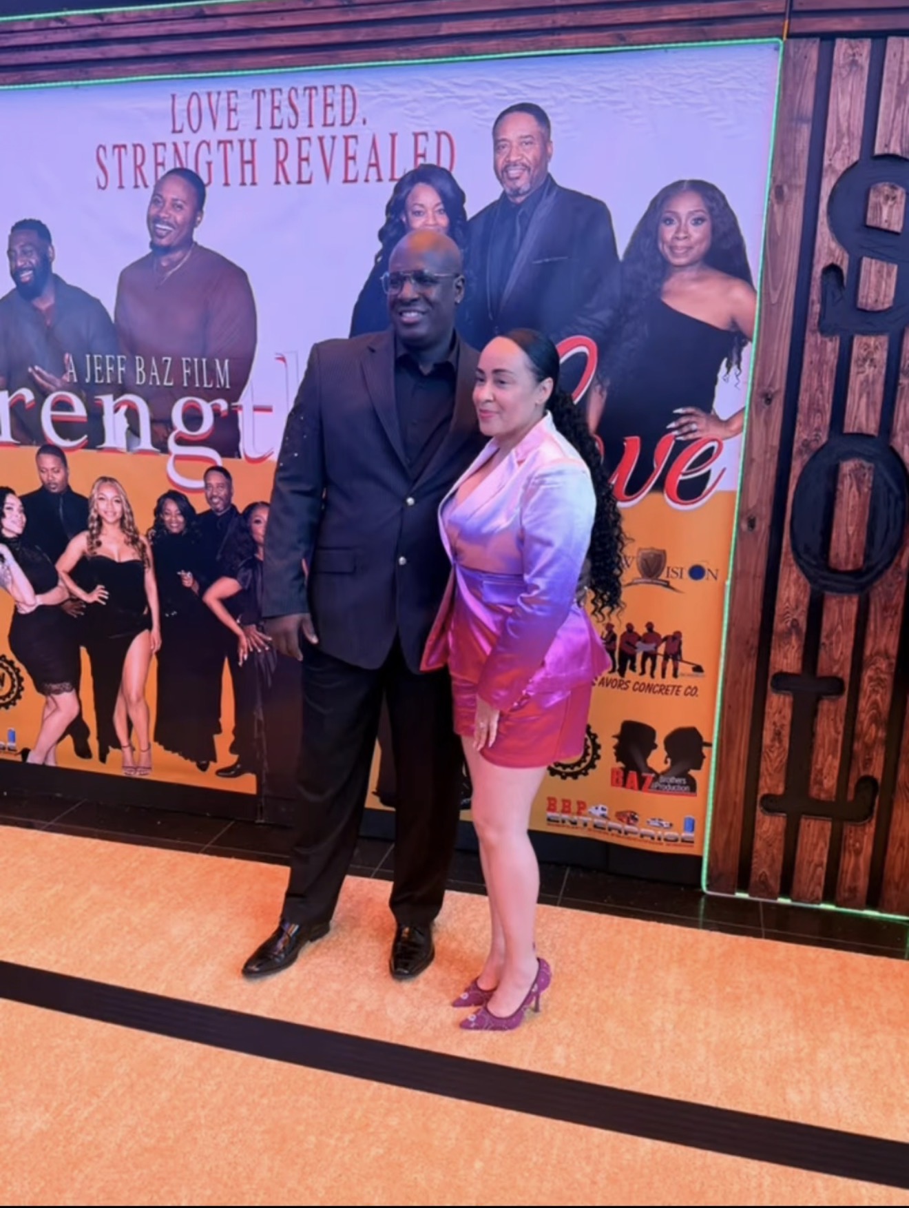 Two people standing in front of a promotional poster at an event, with a group of people in the background.