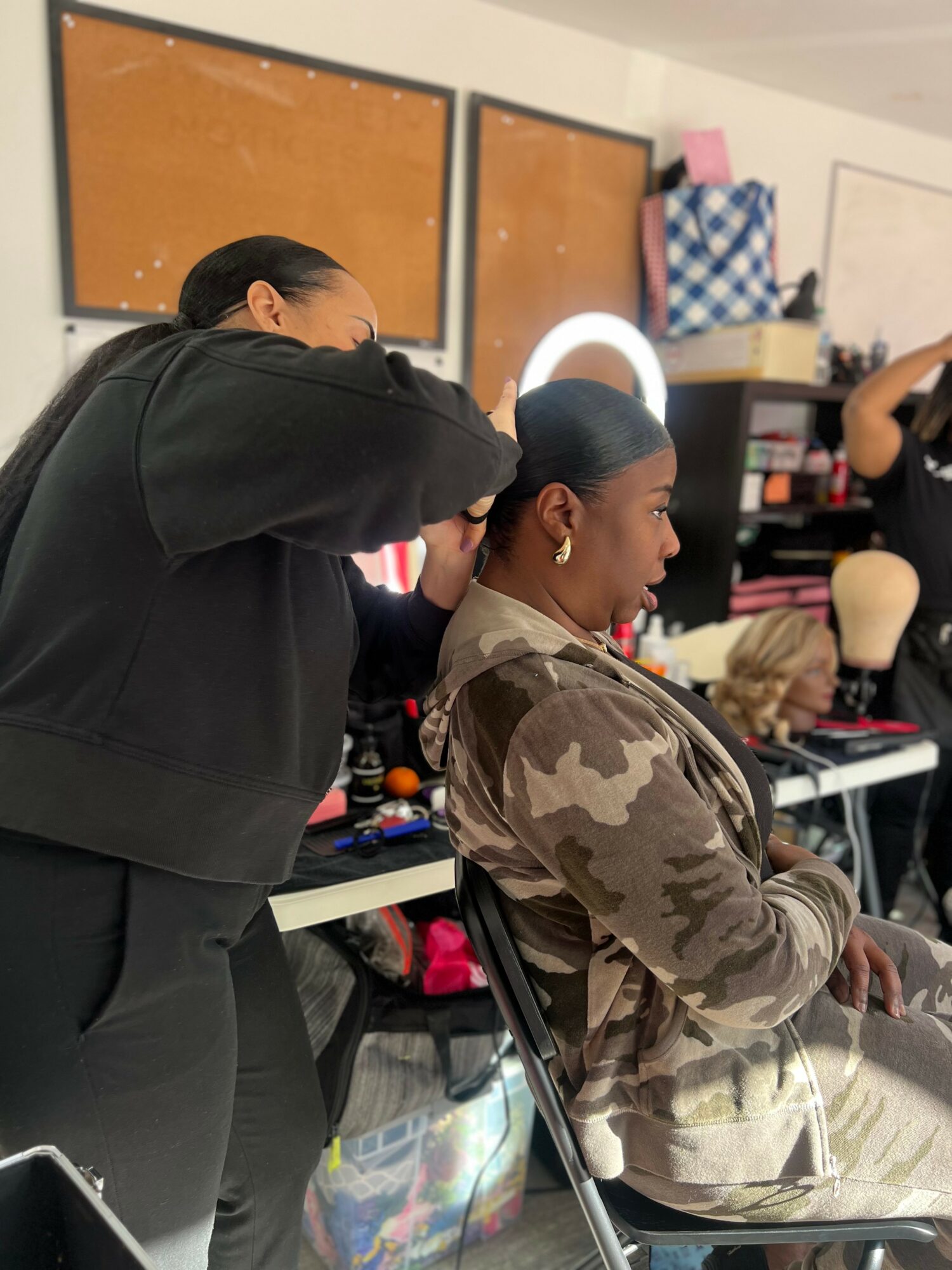 A woman in camouflage clothing sits in a chair while another woman styles her hair in a room with makeup and wig supplies.